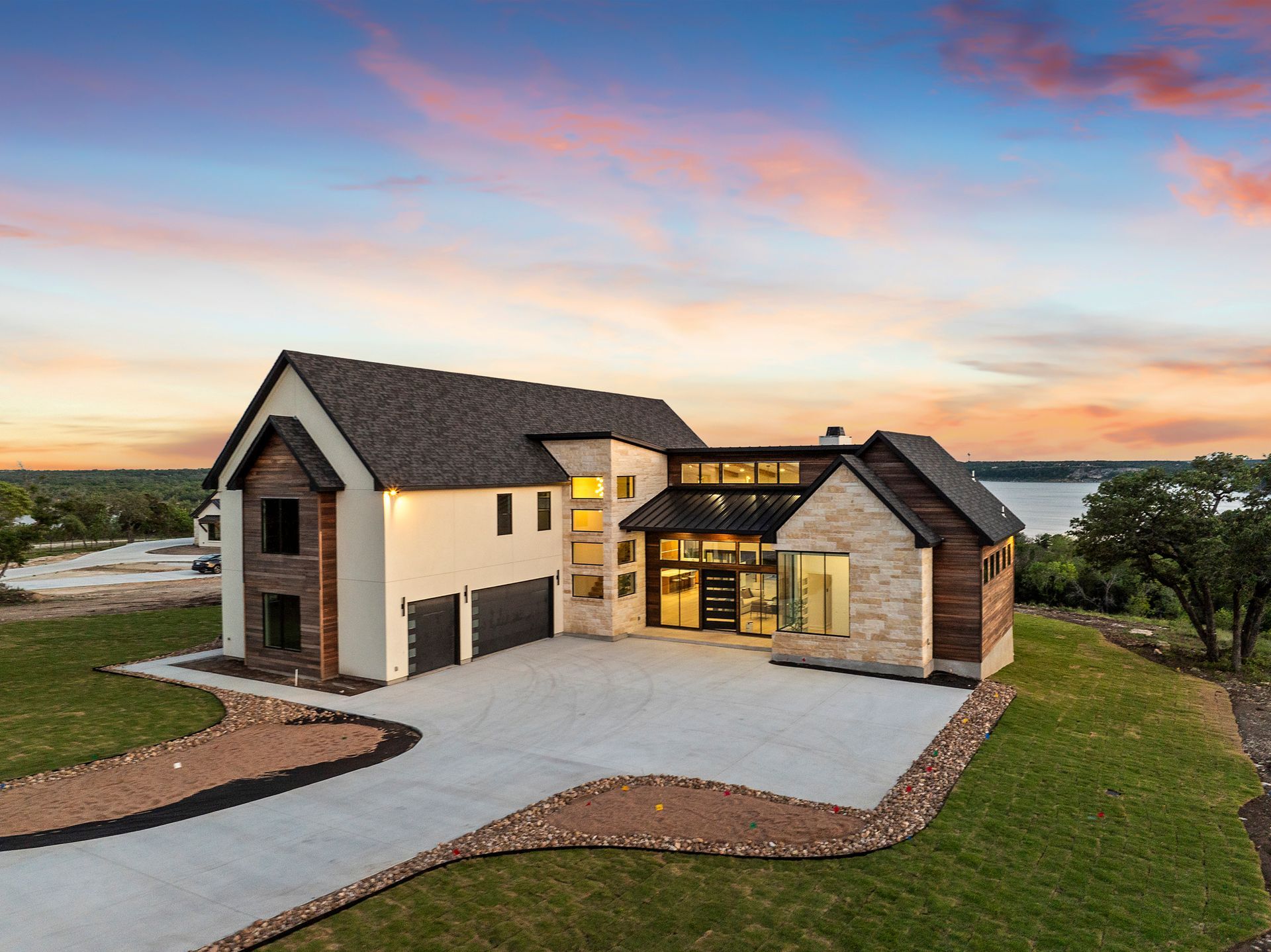 Modern two-story house with lit windows, stone siding, and a curved driveway at sunset