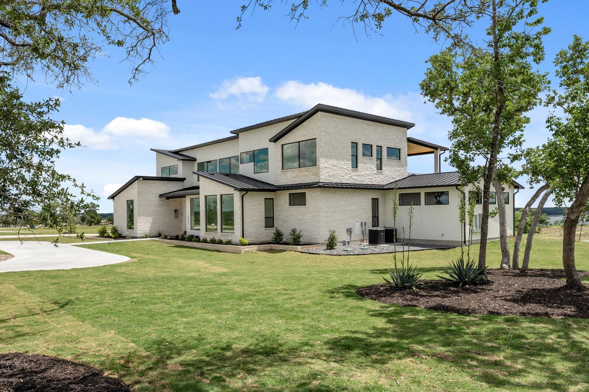 Two-story white house with green roof on a grassy lawn, surrounded by trees under a blue sky.