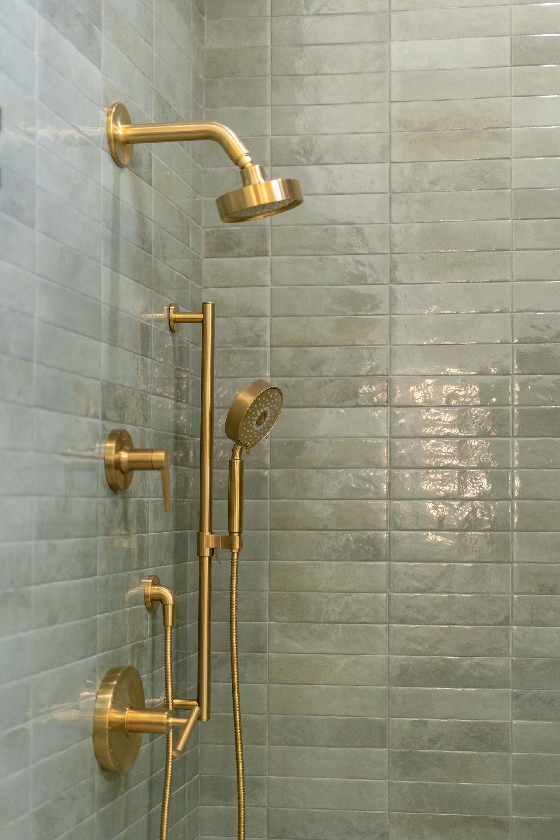 Gold shower fixtures on a tiled shower wall, including an overhead showerhead and handheld sprayer.