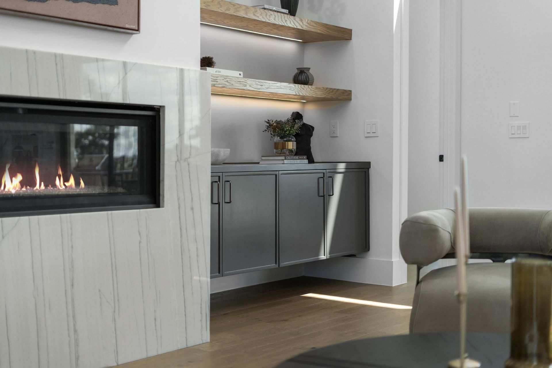 Modern living room with a glass-front fireplace, gray cabinets, open shelves, and a cushioned chair