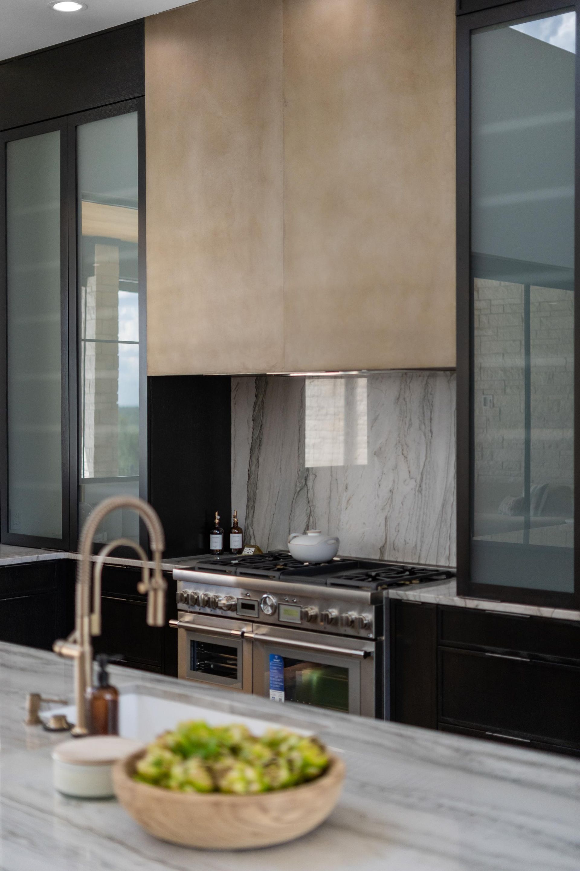 Modern kitchen with marble island, copper faucet, and stainless stove beneath light wood cabinets.