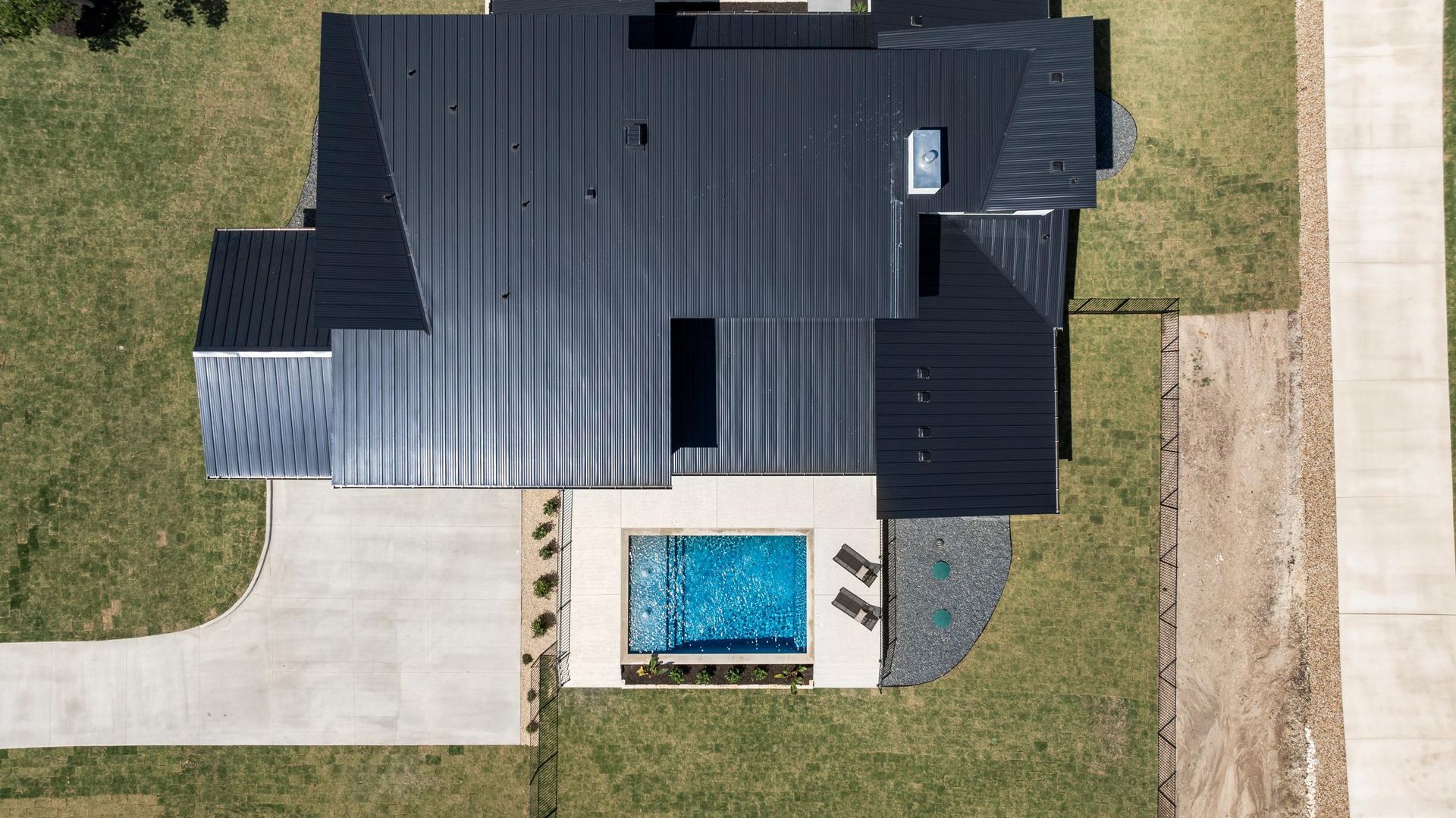 Aerial view of a dark-roofed house with a small backyard pool and driveway beside a dirt road.