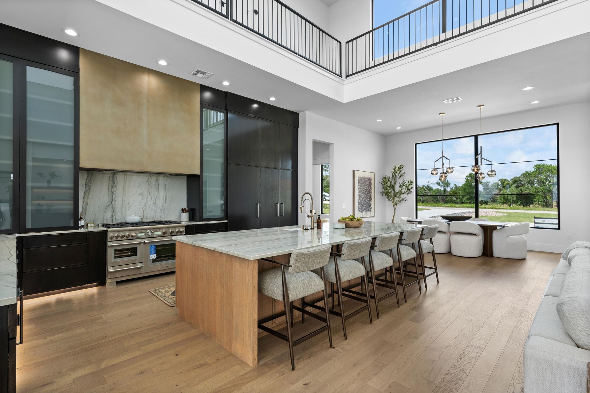 Modern open-plan kitchen and living room with island, wood floors, and large windows overlooking greenery