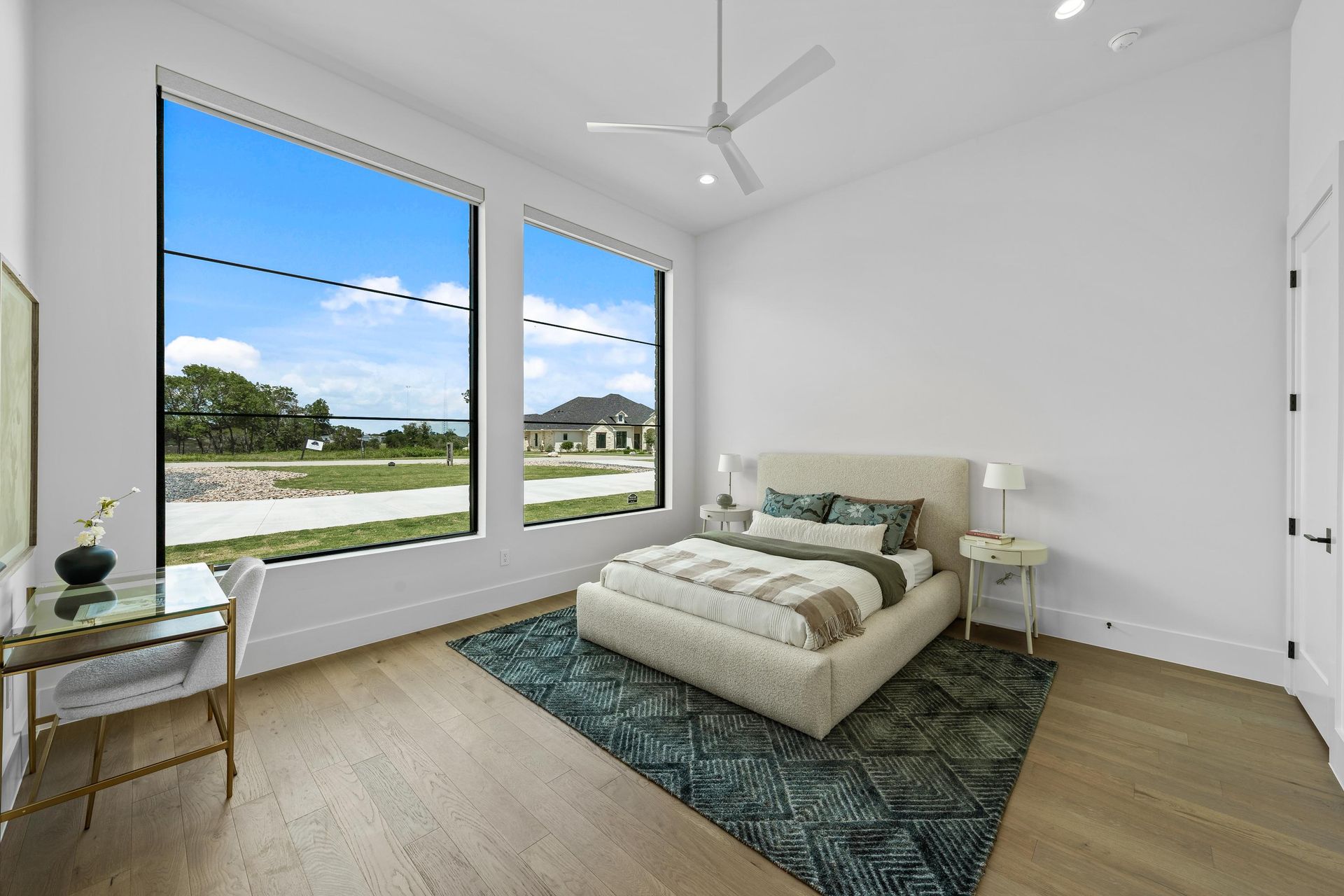 Bright modern bedroom with a bed, rug, and large windows overlooking a grassy neighborhood.