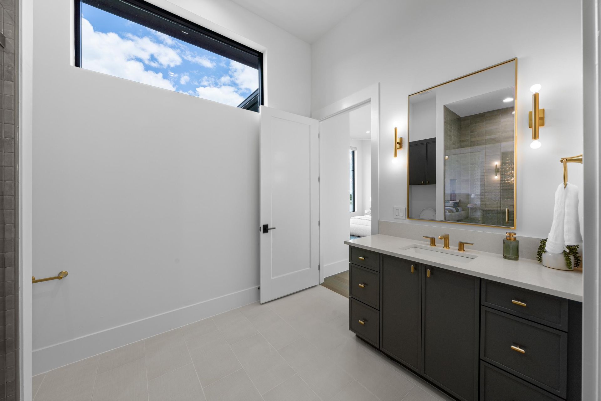 Modern bathroom with dark vanity, large mirror, brass sconces, and a high window.