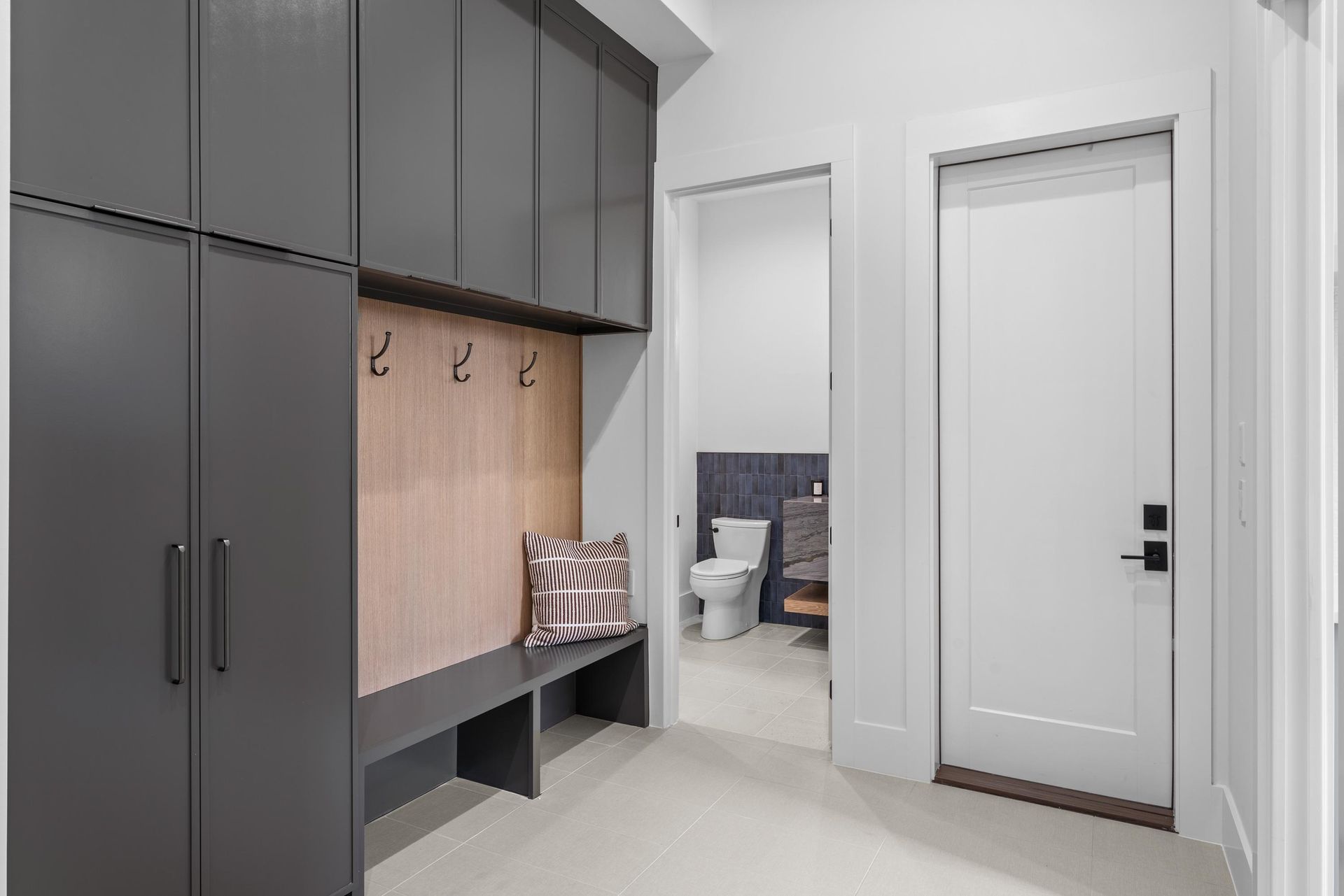 Modern hallway mudroom with dark cabinets, bench, hooks, and a white door to a bathroom.