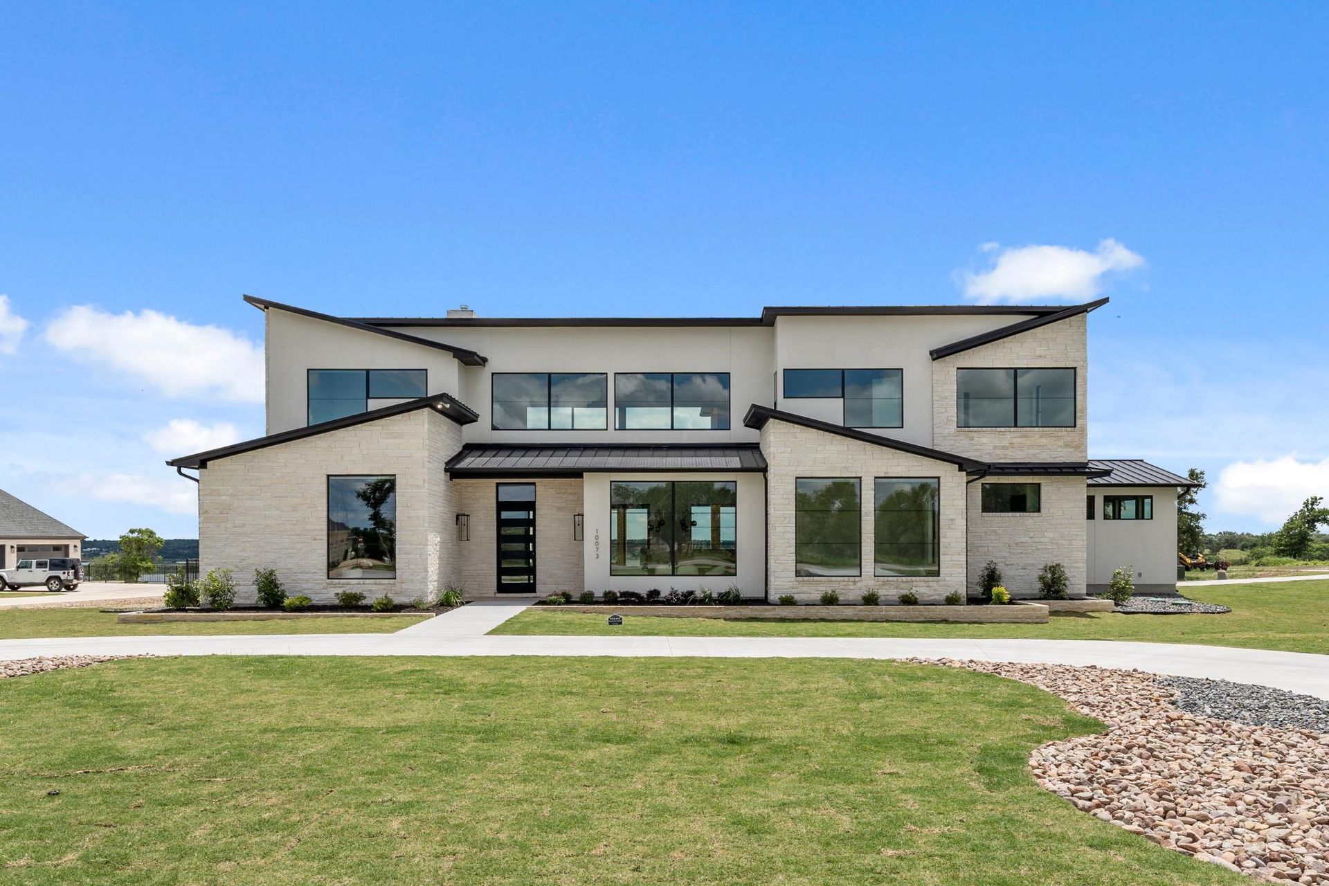 Modern two-story house with beige stone exterior, large windows, and a wide lawn under a blue sky