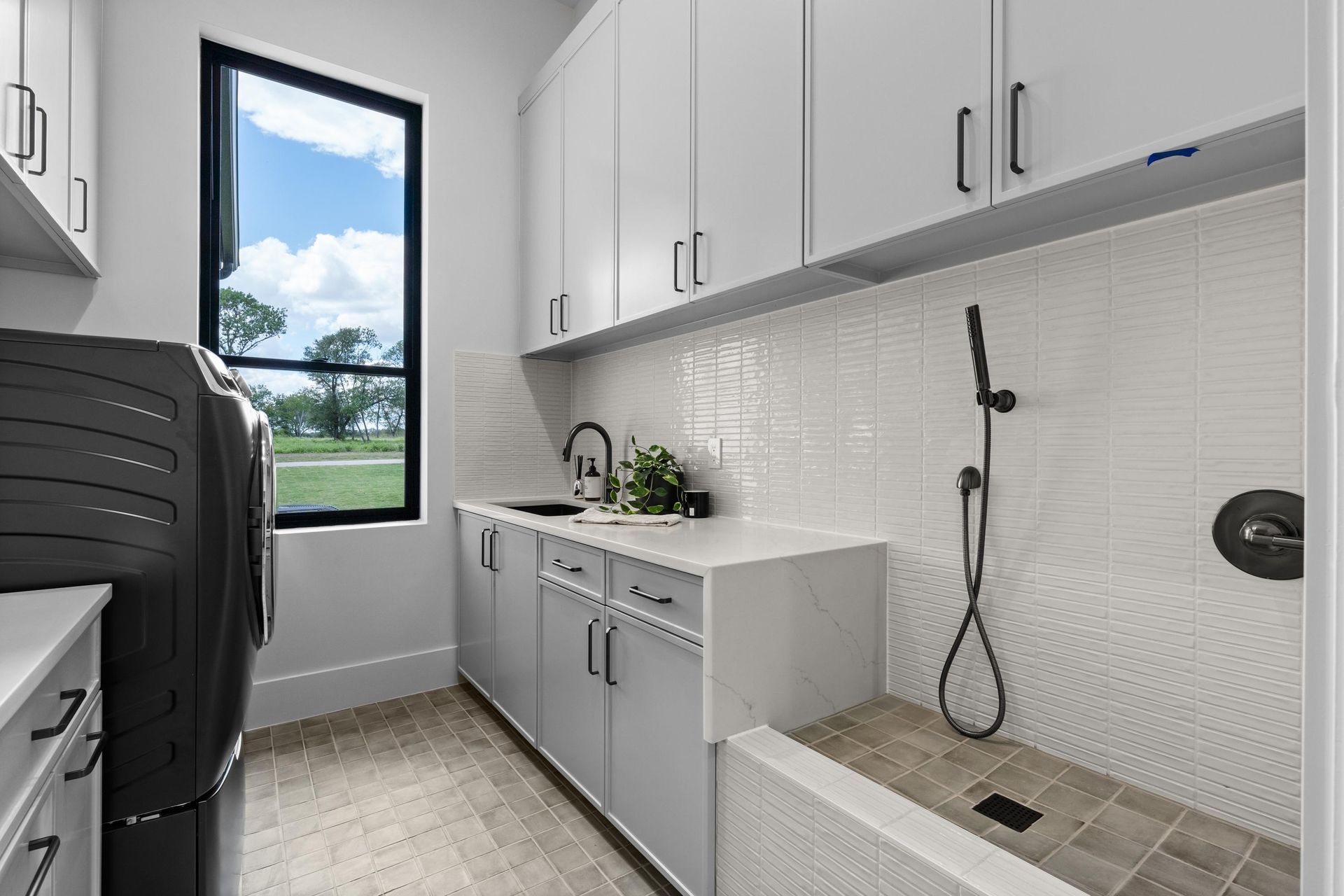 Bright laundry room with white cabinets, sink, washer, and tiled shower area near a window.