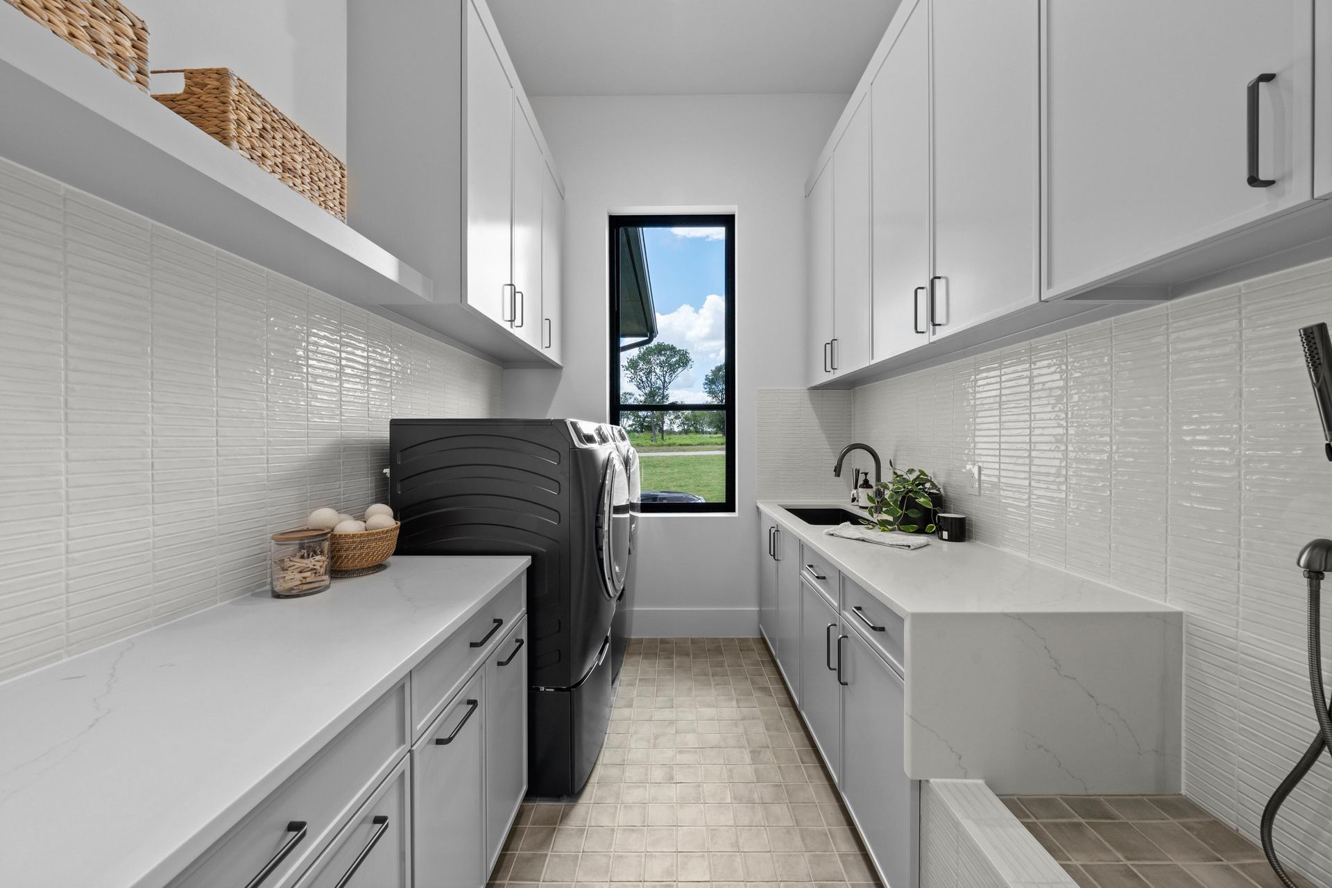 Narrow white galley kitchen with black appliances, gray tile floor, and window at the end.