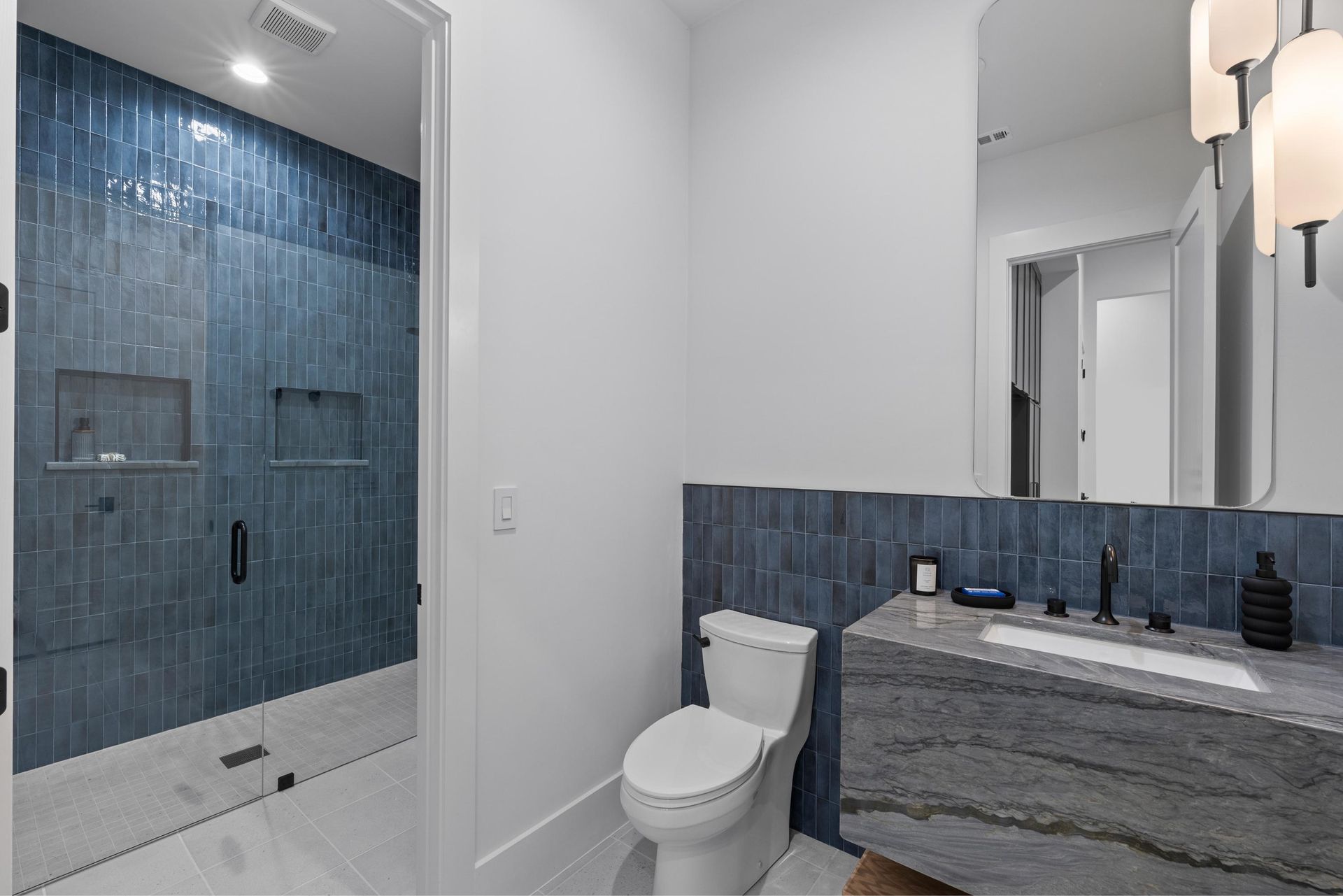 Modern bathroom with blue tiled shower, toilet, and gray vanity with mirror and lights