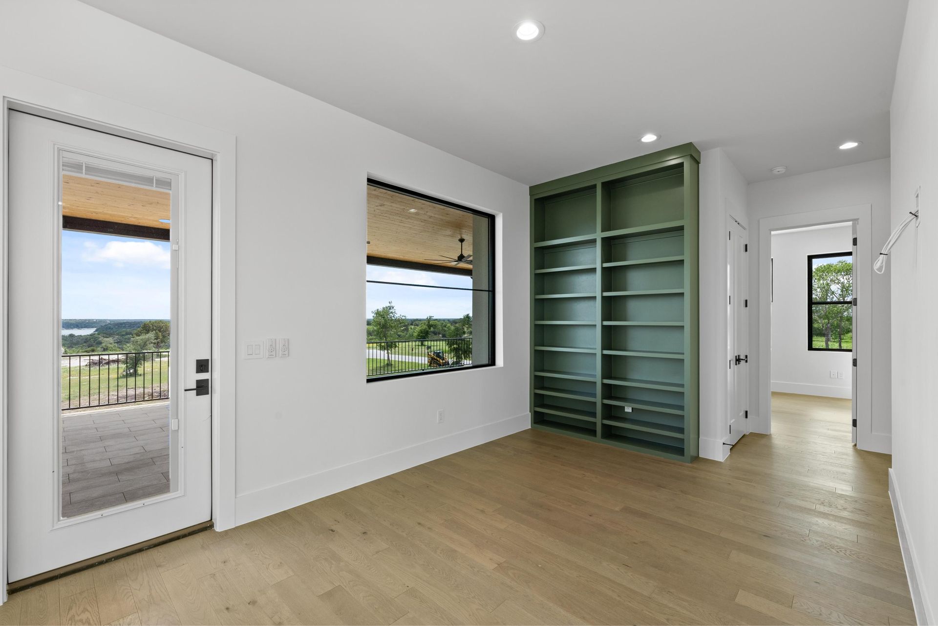 Empty modern room with light wood floors, white walls, green built-in shelves, and balcony view through windows