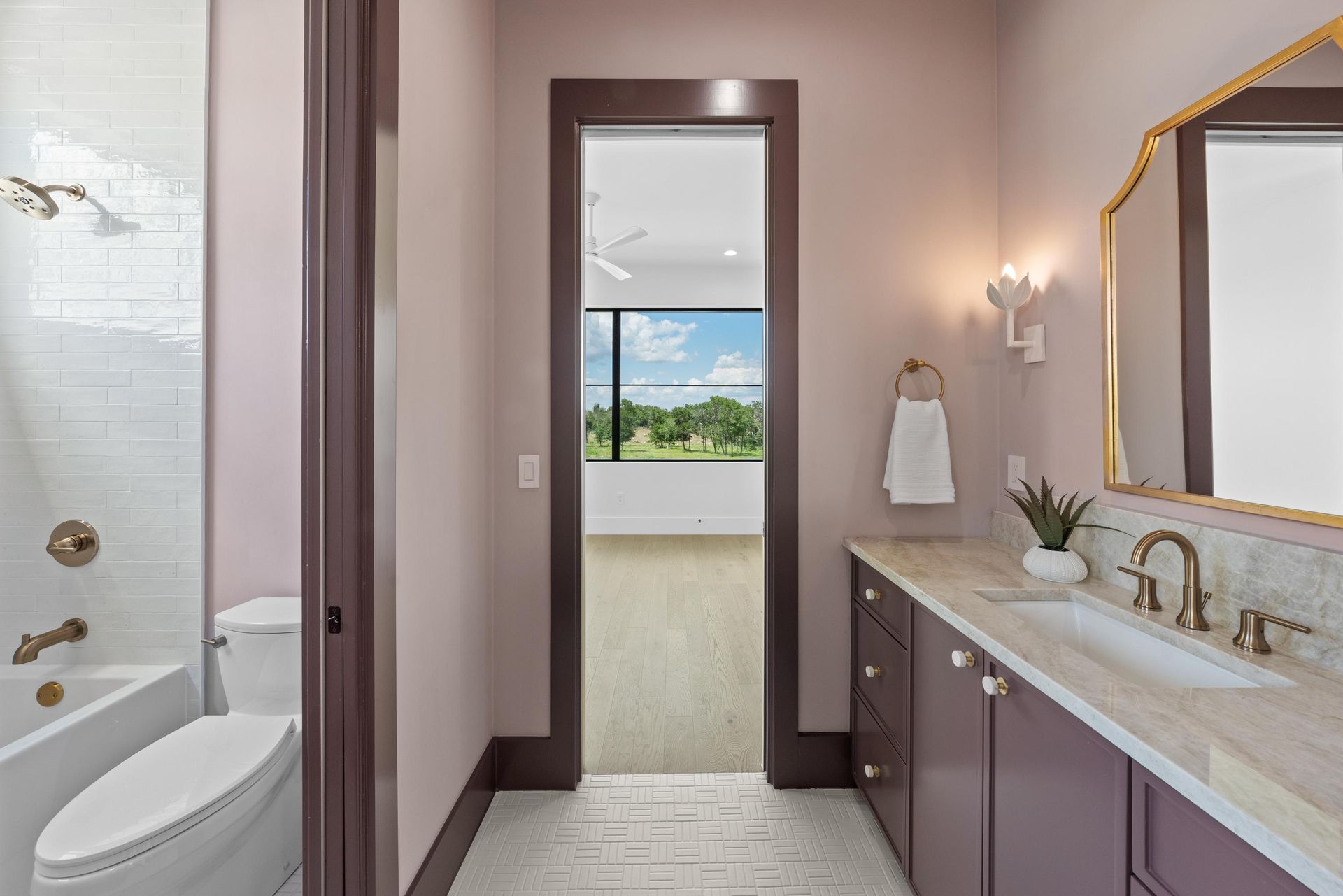 Modern bathroom with dual-sink vanity, marble counter, and open doorway to a bright room with green views
