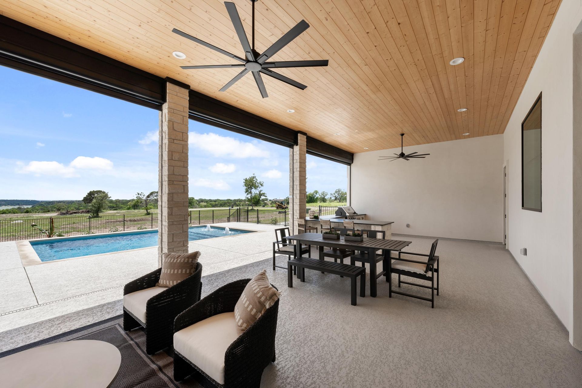 Covered patio with dining set and lounge chairs beside a pool, overlooking open countryside.