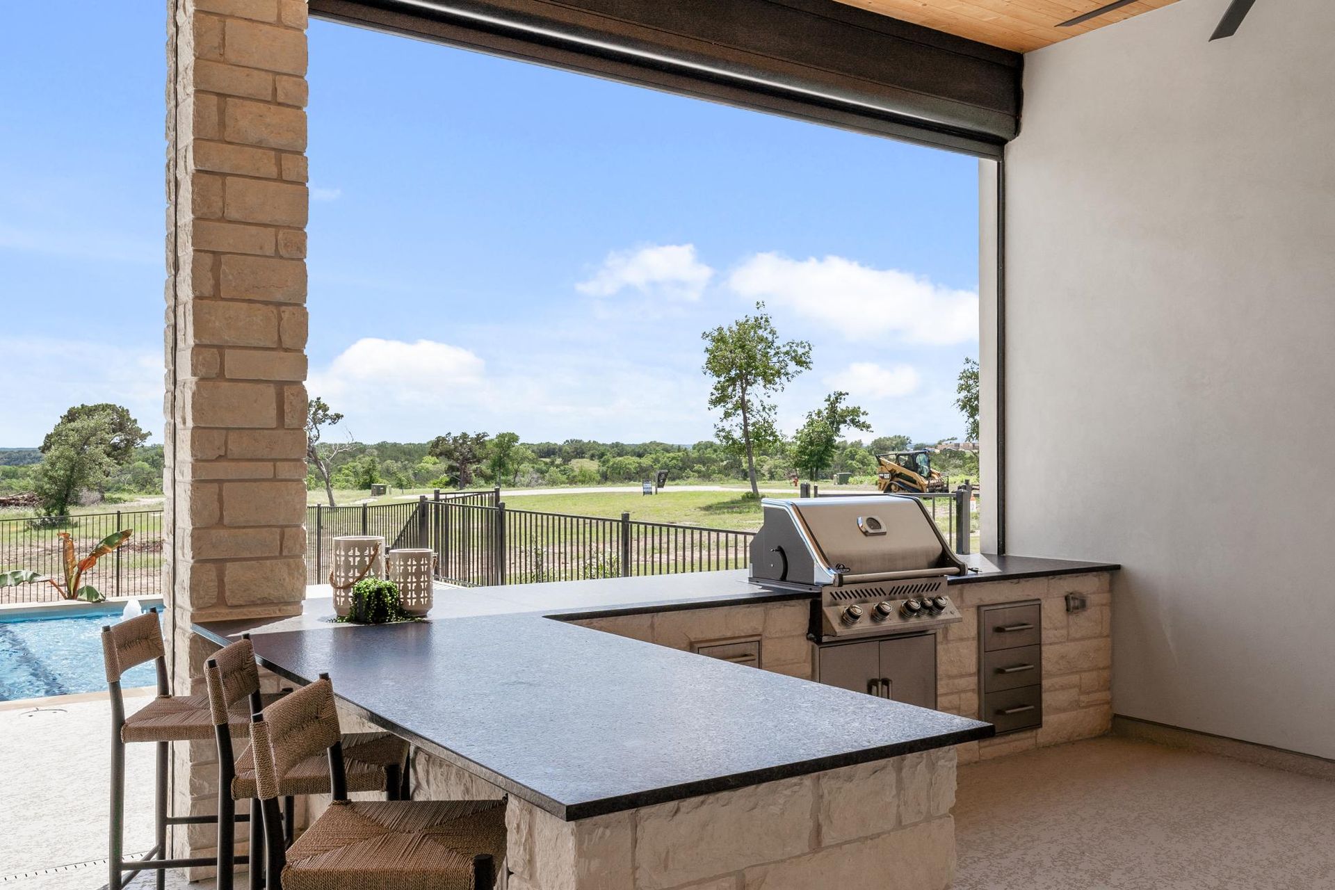 Covered patio with outdoor kitchen, dining table, and open view of pool and green fields