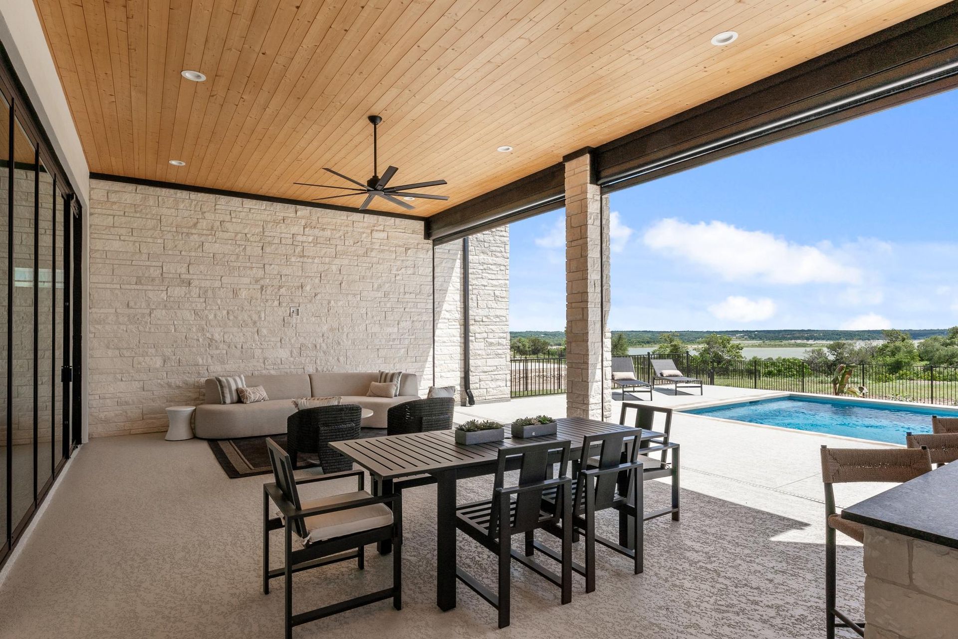 Covered patio dining area with black chairs, pool, and ocean view under a wood سقiling
