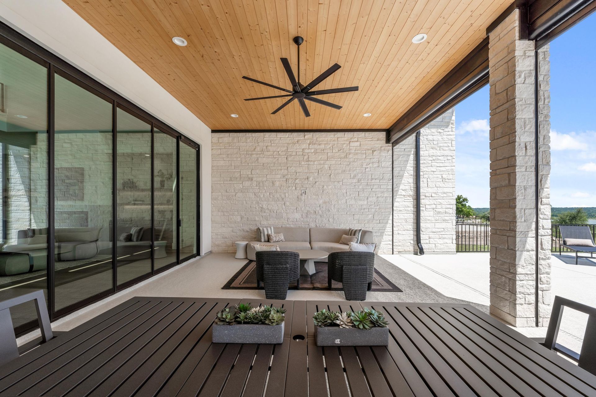 Covered patio with wood ceiling, black ceiling fan, glass walls, and outdoor seating on a deck.