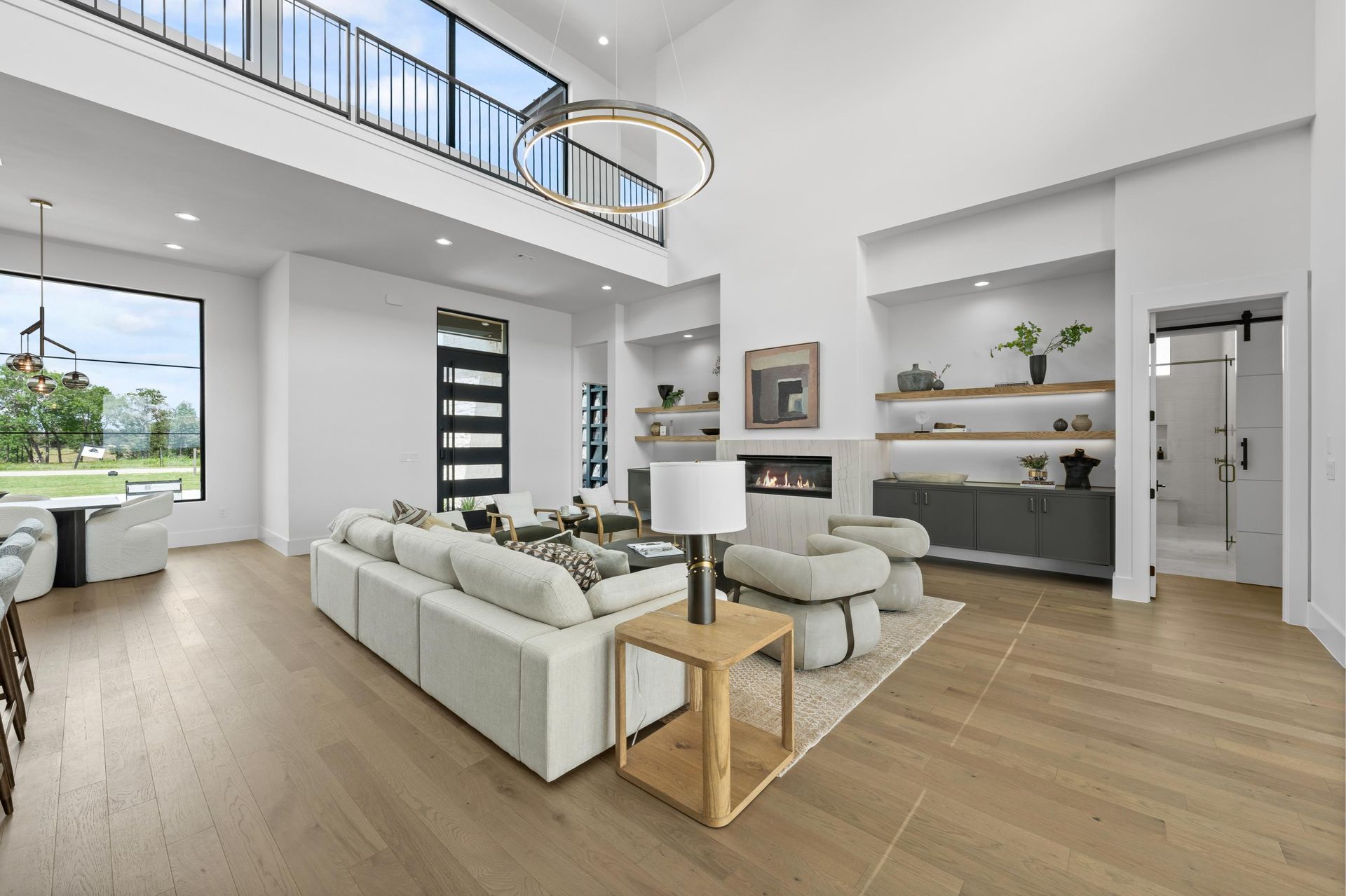 Bright modern living room with white sofas, wood floors, tall windows, and an open staircase.