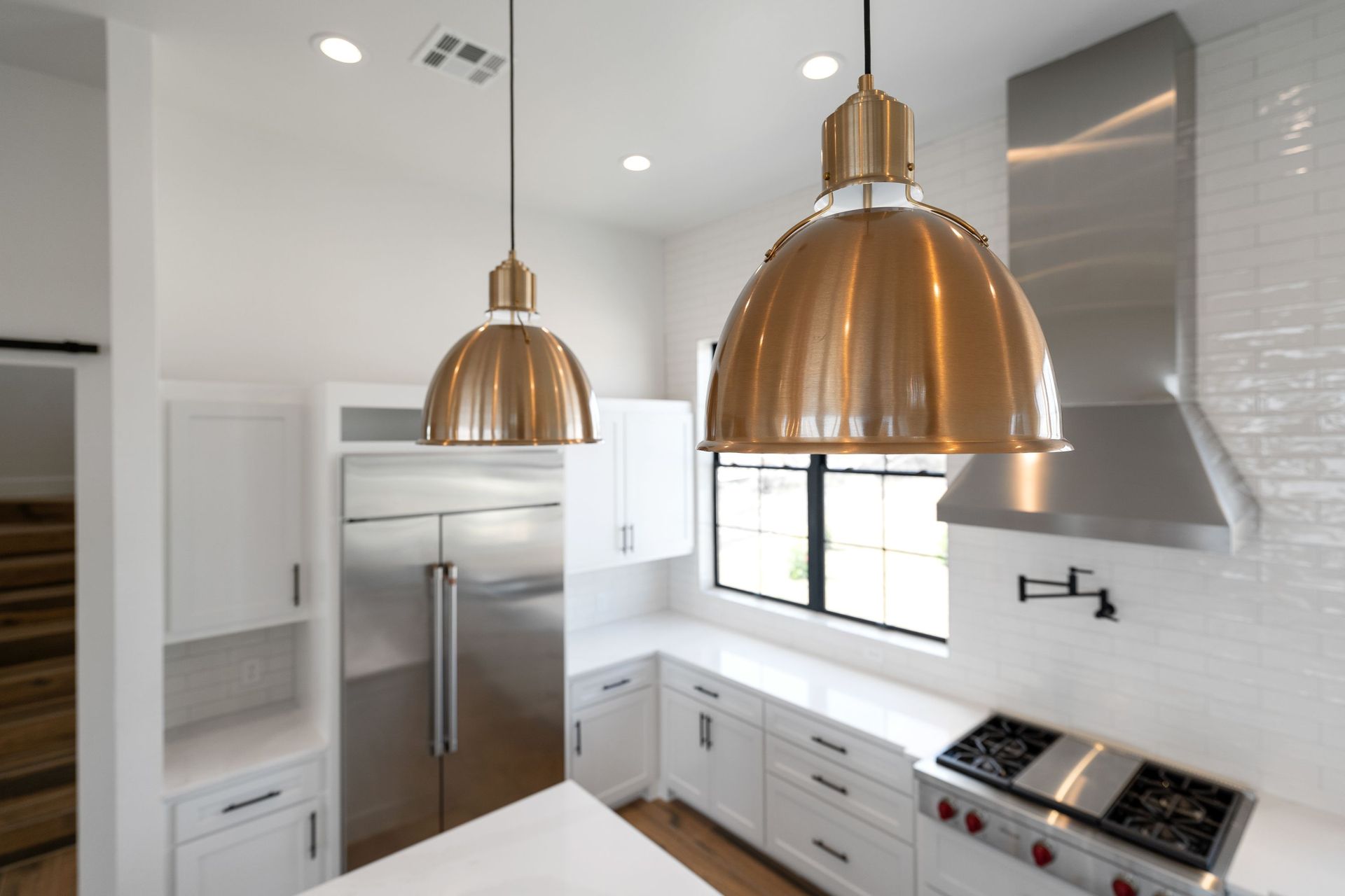 Modern white kitchen with stainless steel fridge, brass pendant lights, and large window above a cooktop
