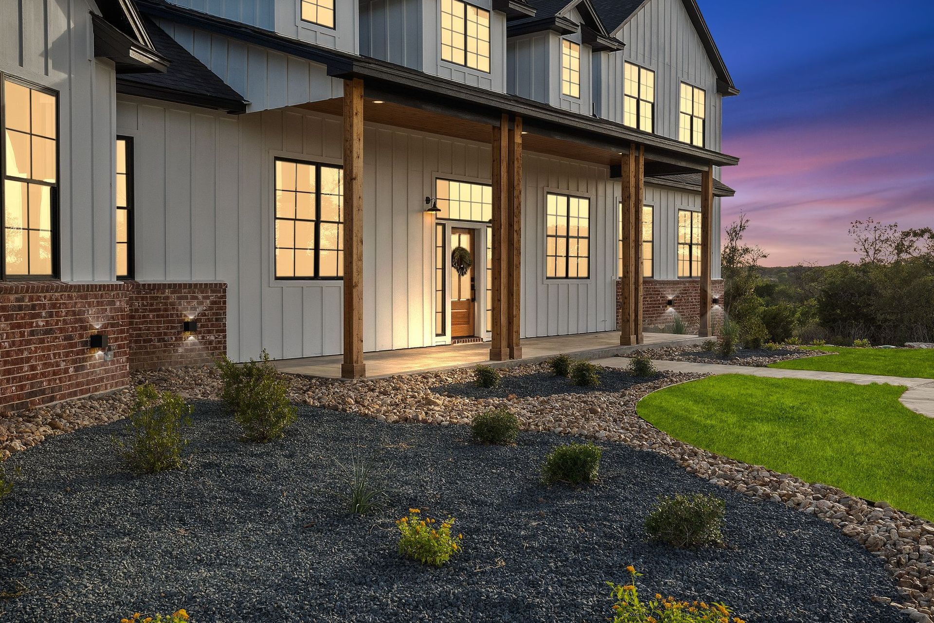 Large modern house exterior at dusk with lit windows, front porch, and landscaped gravel and lawn yard.