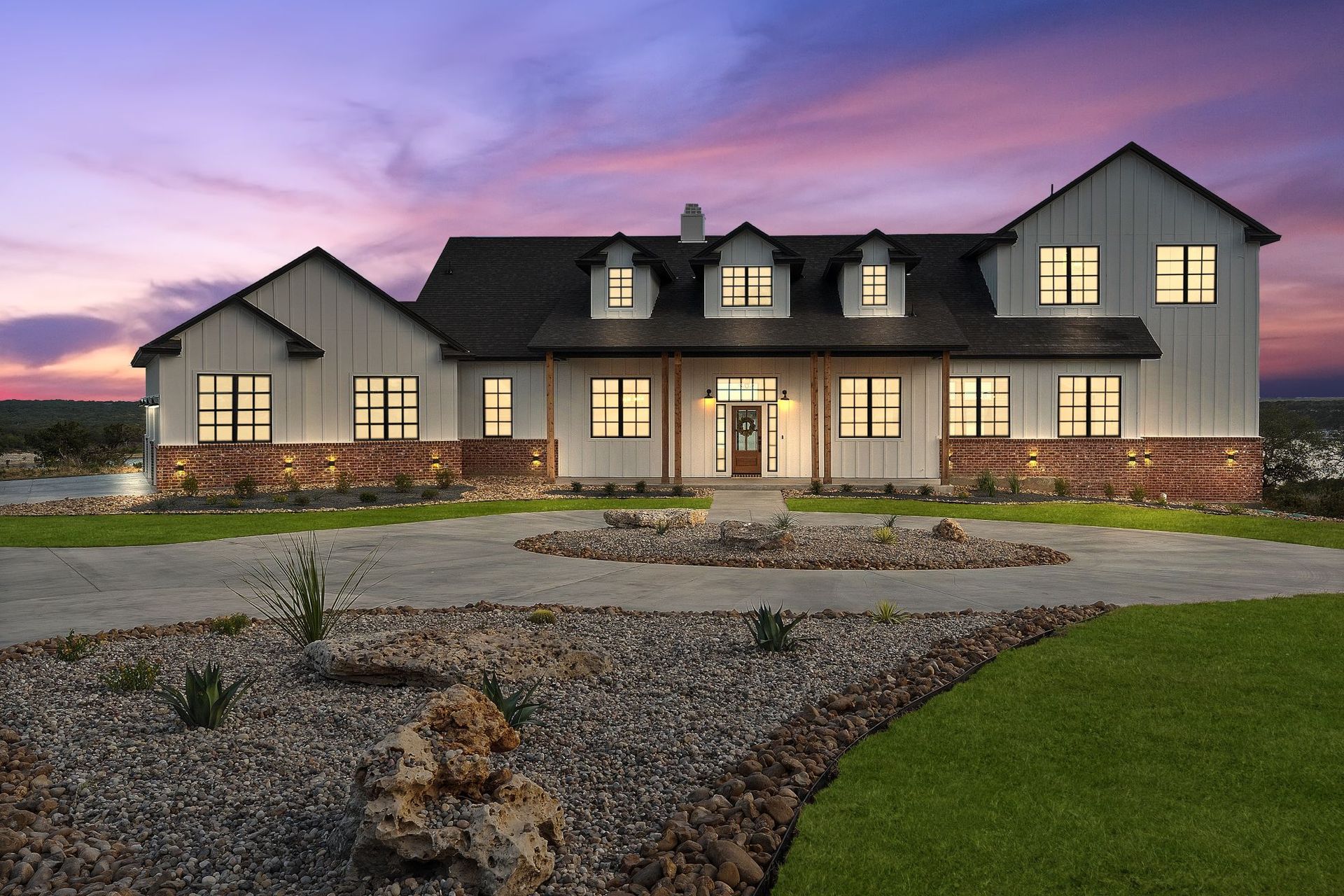 Large gray suburban house at dusk with a lit porch, stone accents, and landscaped front yard
