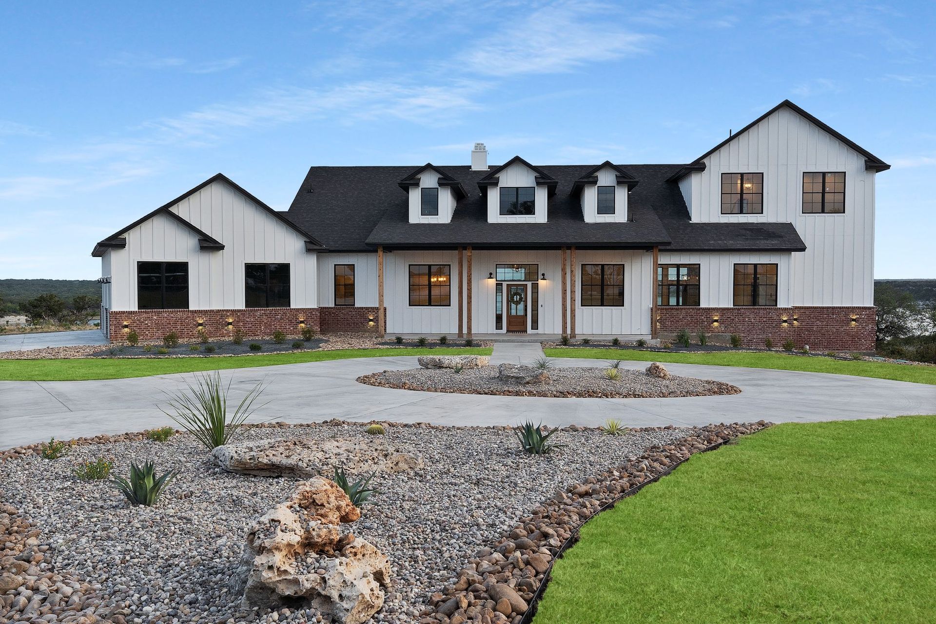 Large white farmhouse with black roof, stone accents, and a landscaped gravel driveway under a cloudy sky