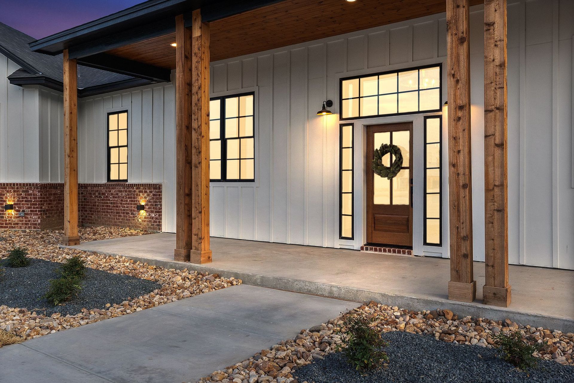 Front porch of a house with lit windows, wooden posts, and a wreath on the front door at dusk