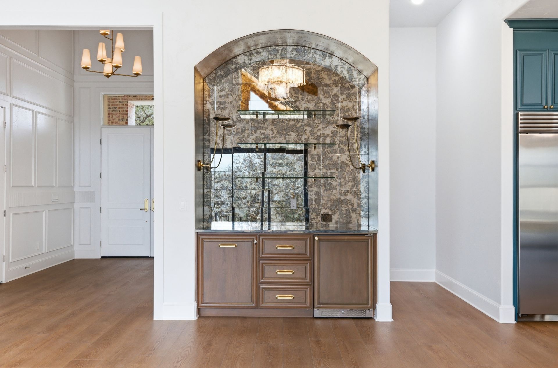Empty room with wood floors, white walls, arched mirrored cabinet, and partial kitchen view