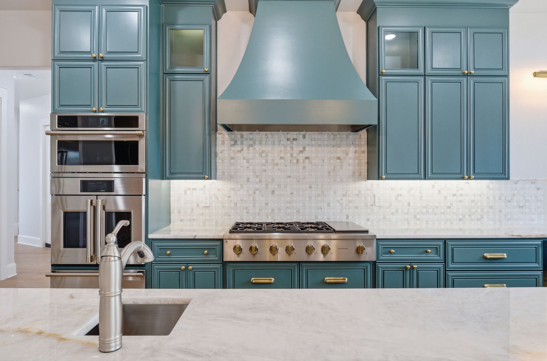 Blue kitchen with stainless appliances, large range hood, and marble island with faucet