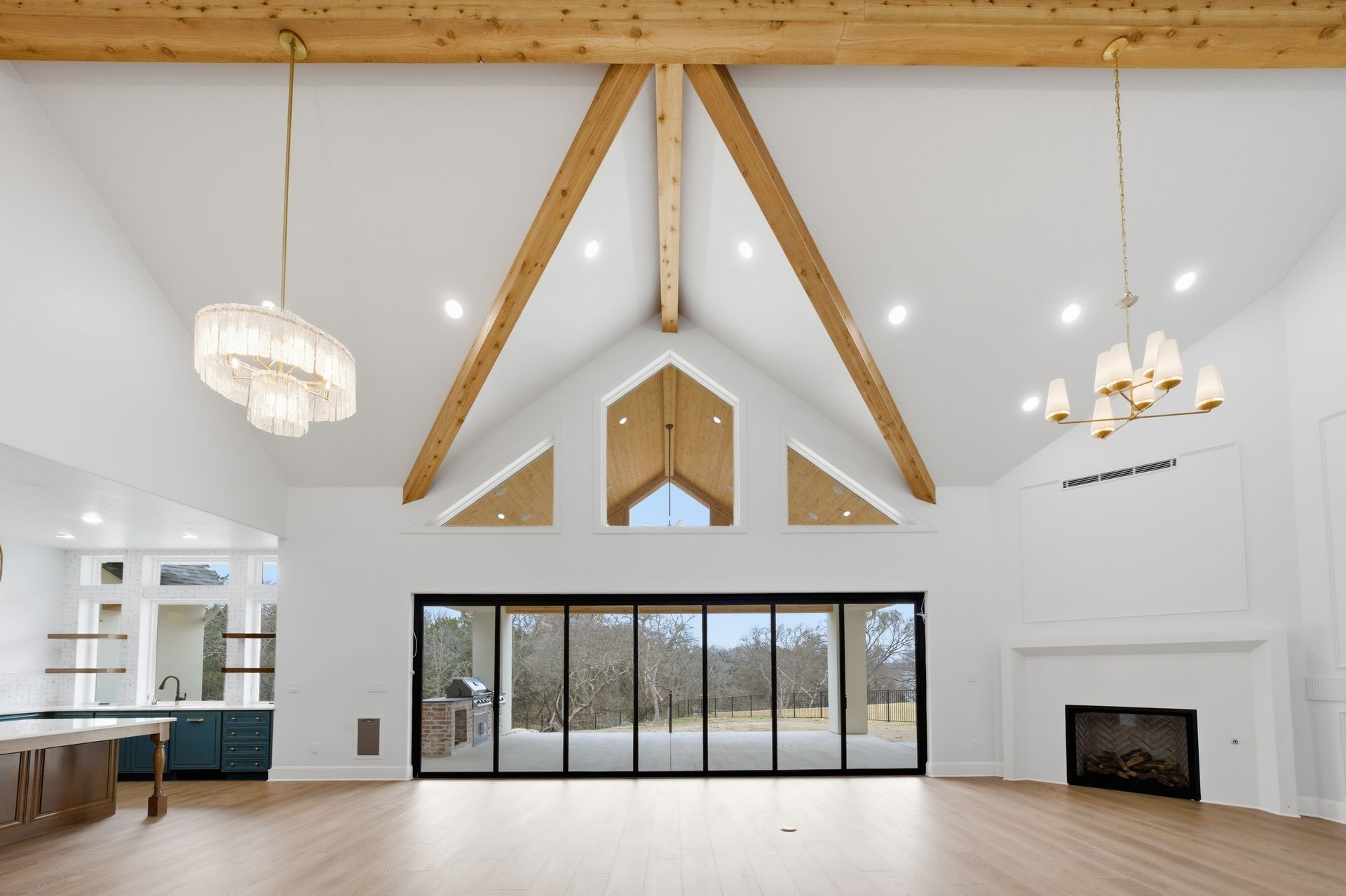 Bright vaulted living room with exposed wood beams, large windows, fireplace, and kitchen view