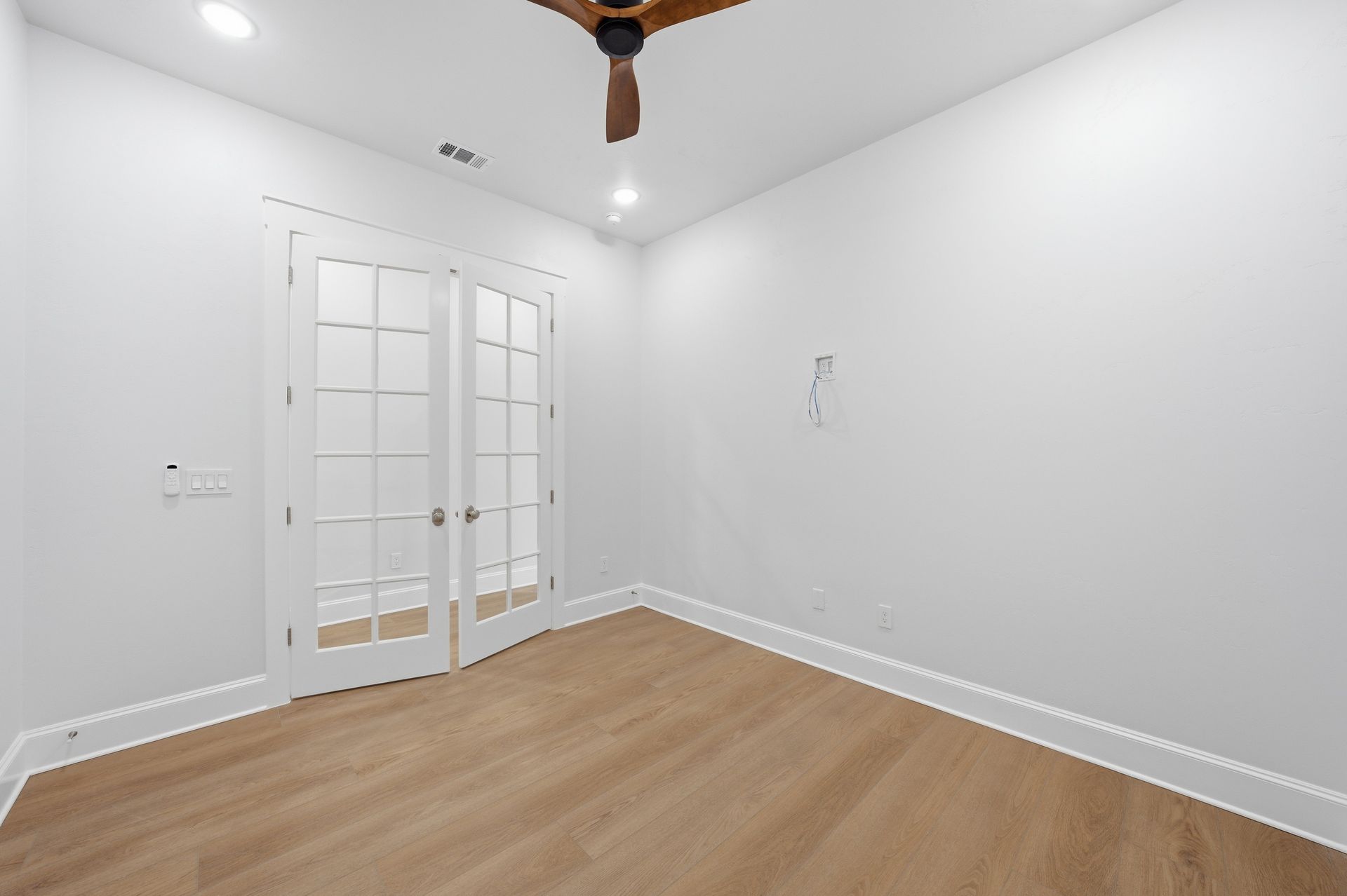 Empty white room with light wood flooring, French doors, and a ceiling fan.