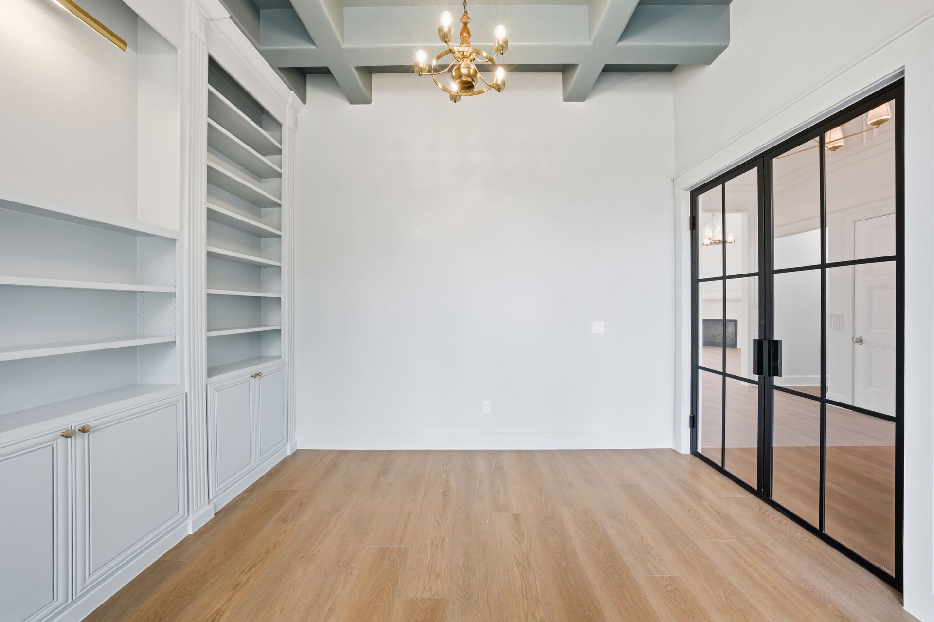 Bright empty room with built-in white shelves, wood floor, chandelier, and glass doors.