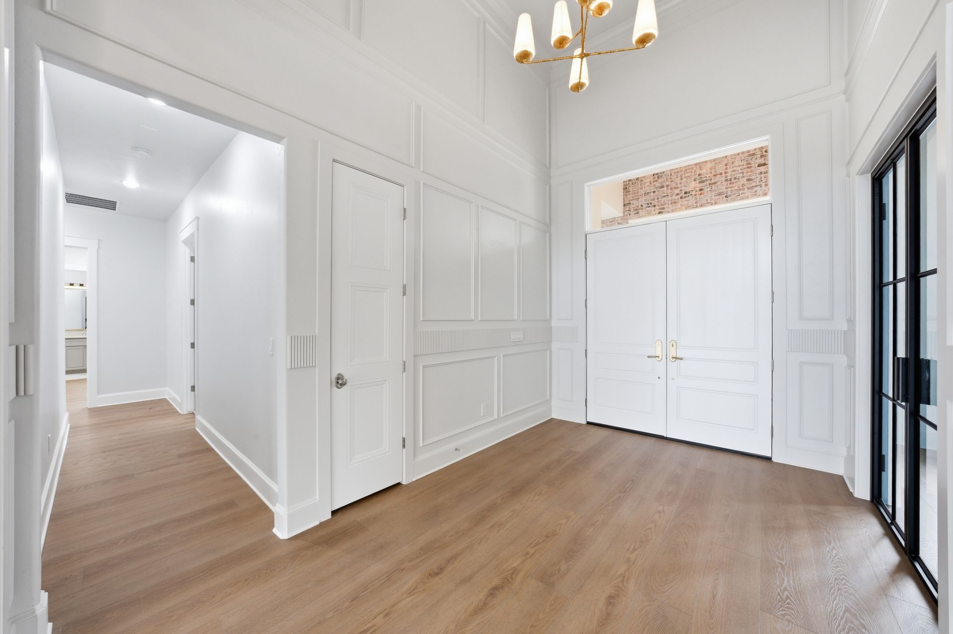 Bright empty entry hall with white paneled walls, wood floors, chandelier, and black-framed glass doors