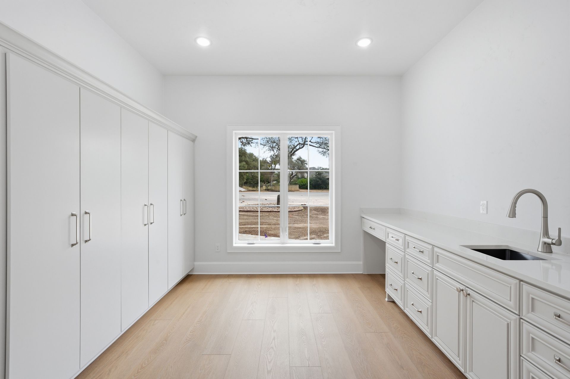 Bright empty laundry room with white cabinets, sink, wood floor, and a window.