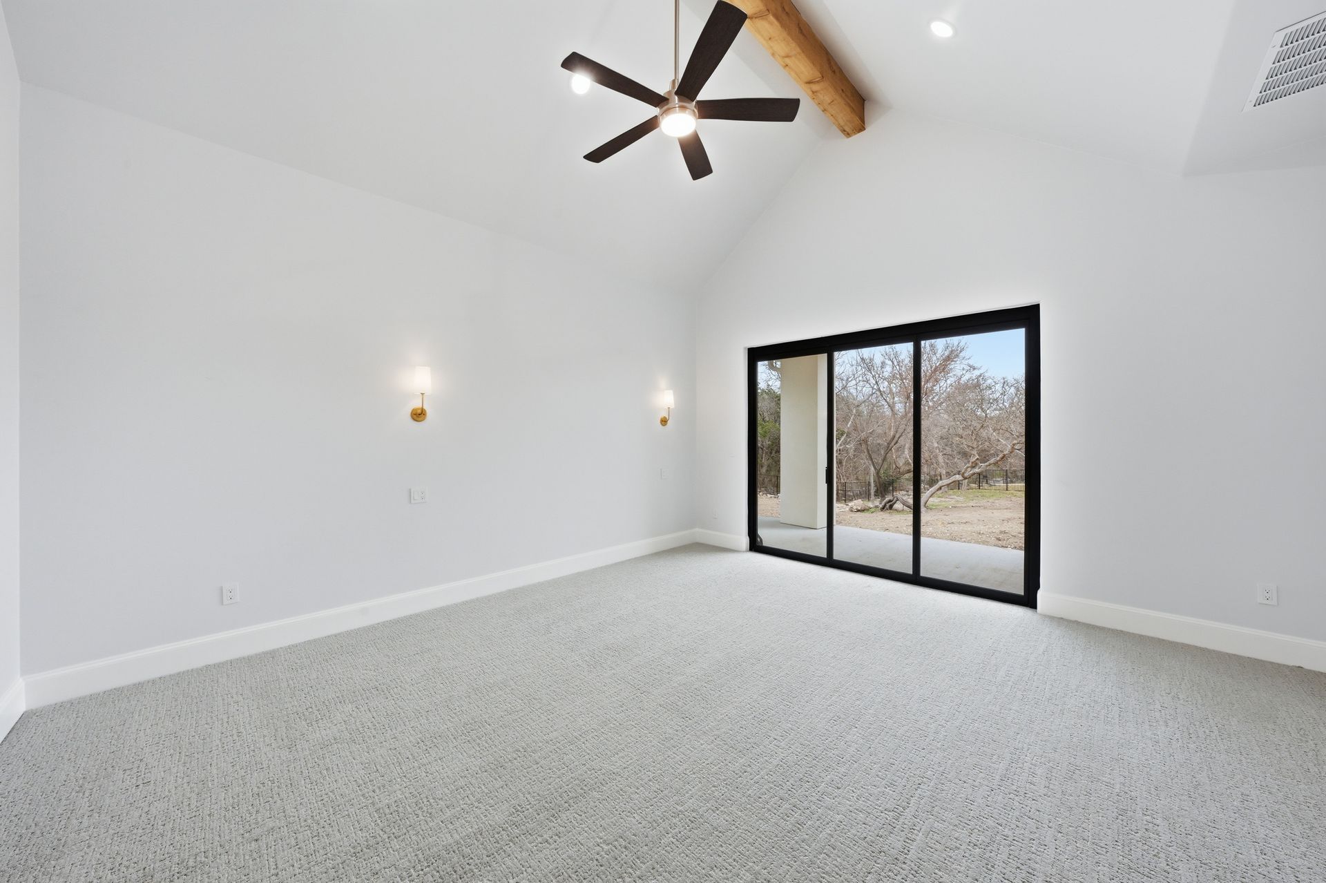 Empty white room with vaulted ceiling, ceiling fan, wall sconces, and large sliding glass doors.