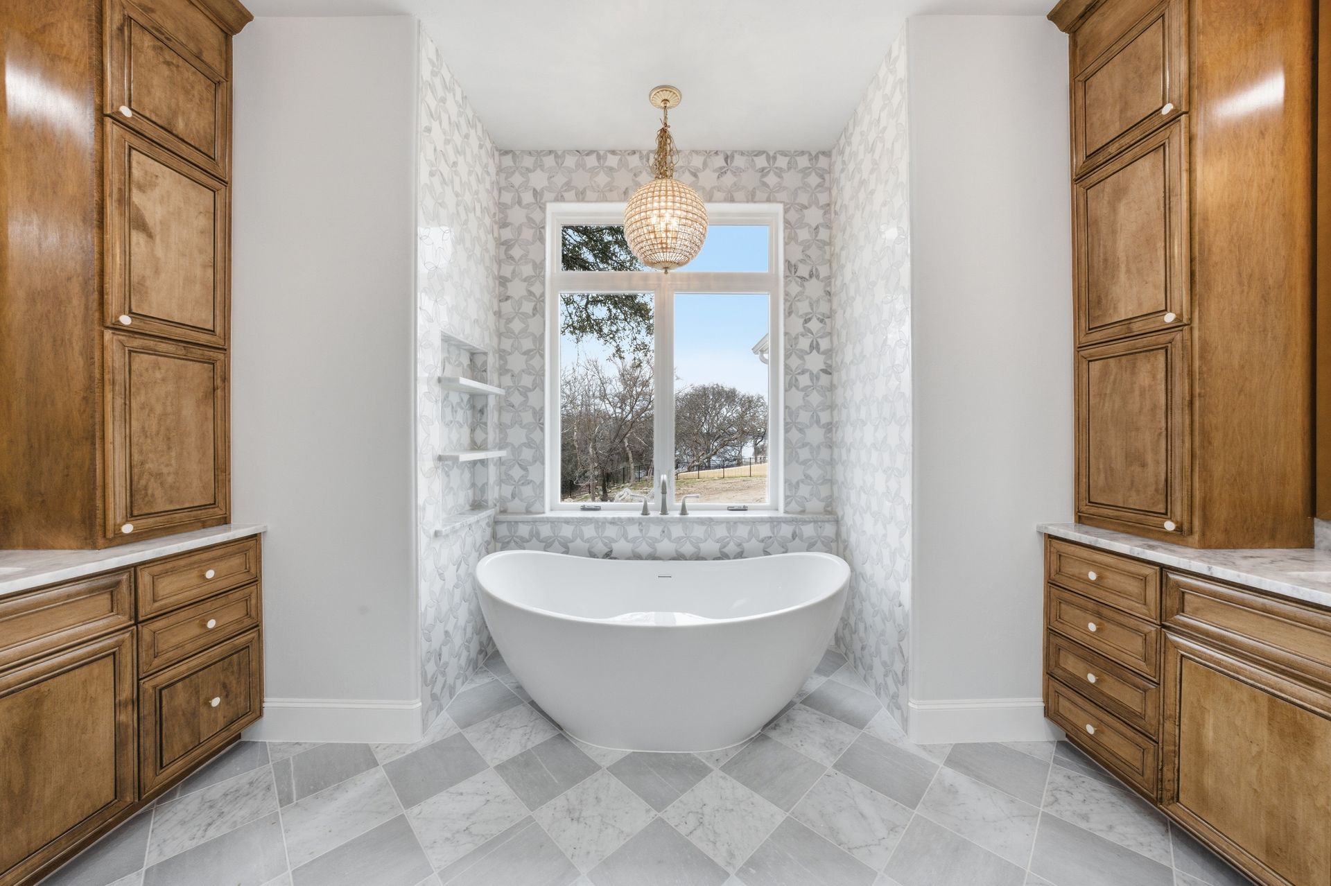 Bright bathroom with a freestanding tub, wooden cabinets, patterned tile floor, and a window centered above the tub