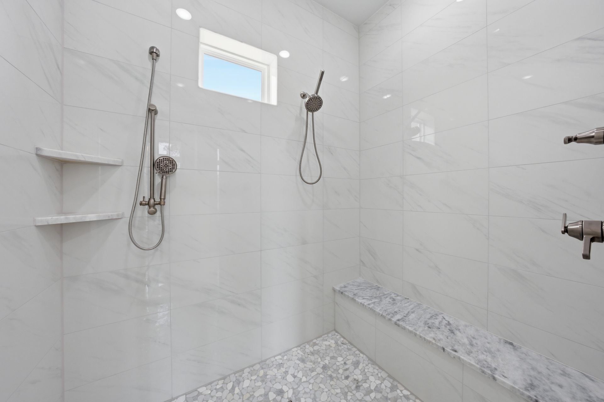 White tiled walk-in shower with multiple showerheads, built-in shelf, and pebble floor.
