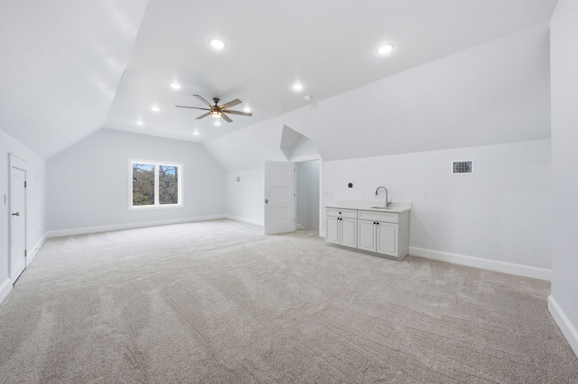 Bright empty carpeted room with white walls, recessed lights, ceiling fan, and small kitchenette area