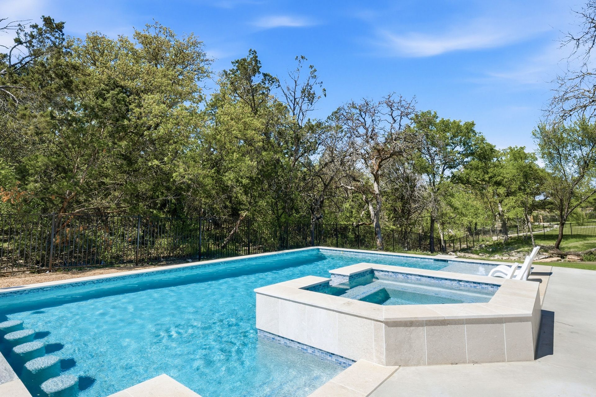 Outdoor swimming pool with a hot tub, white deck, and trees under a blue sky