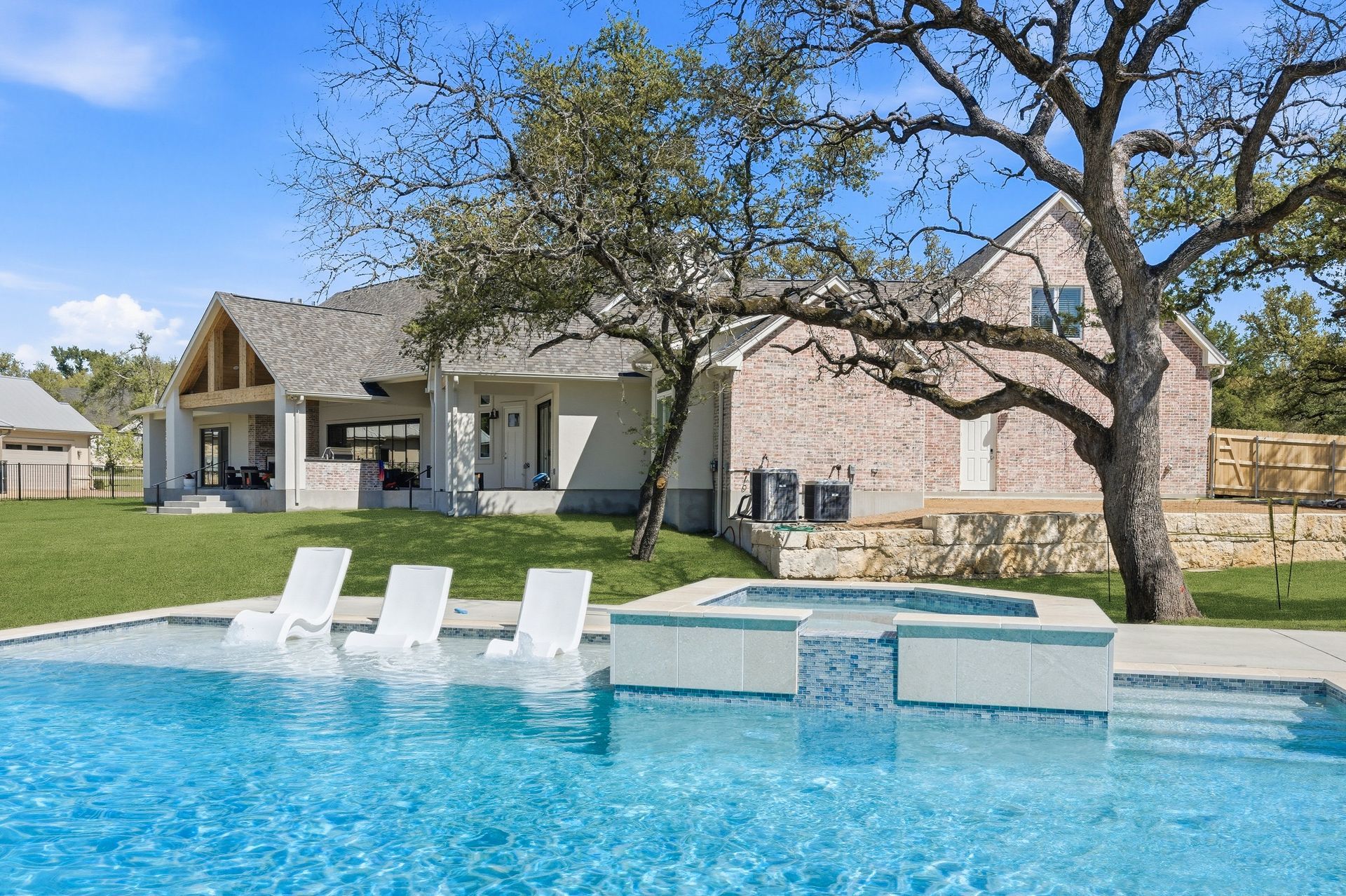 Backyard pool with white lounge chairs beside a brick house and large tree under a blue sky