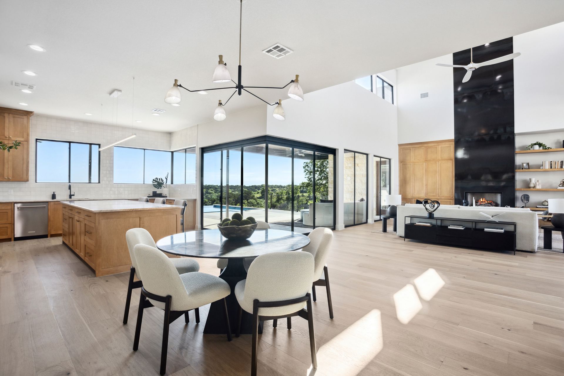 Modern open-concept living room with dining table, kitchen island, and large sliding glass doors overlooking greenery
