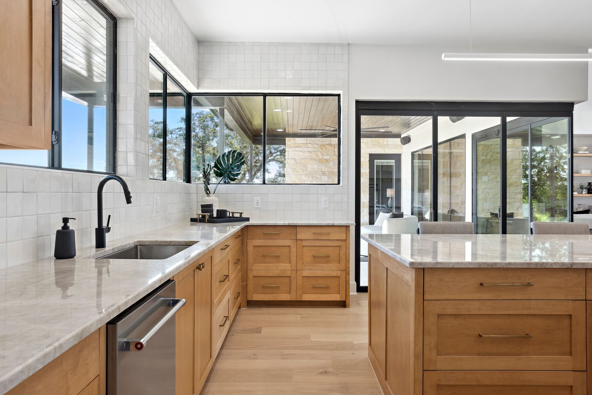 Bright modern kitchen with white counters, wood cabinets, black fixtures, and large windows.