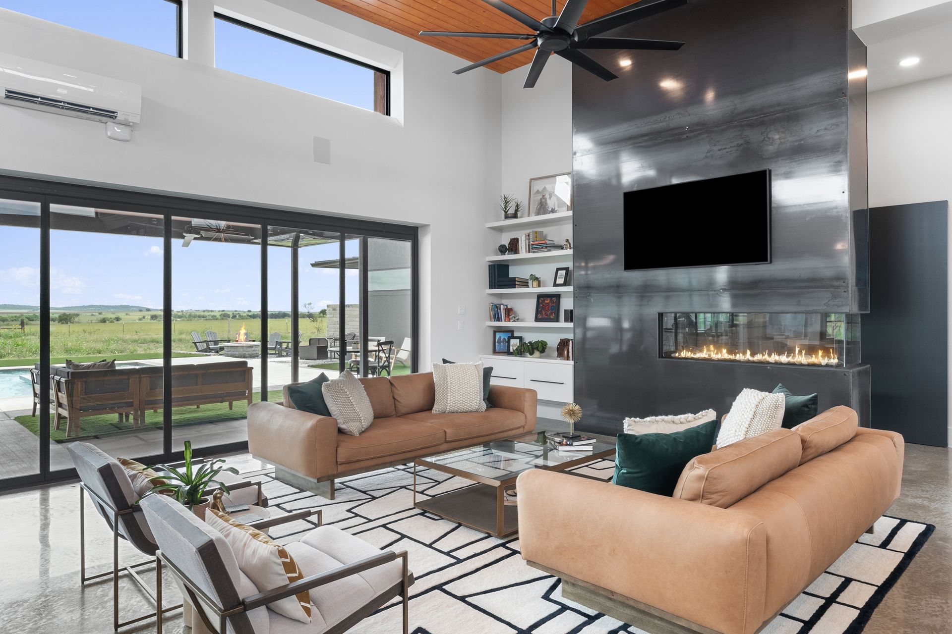 Modern living room with tan sofas, fireplace TV, large windows, and a high ceiling with ceiling fan.