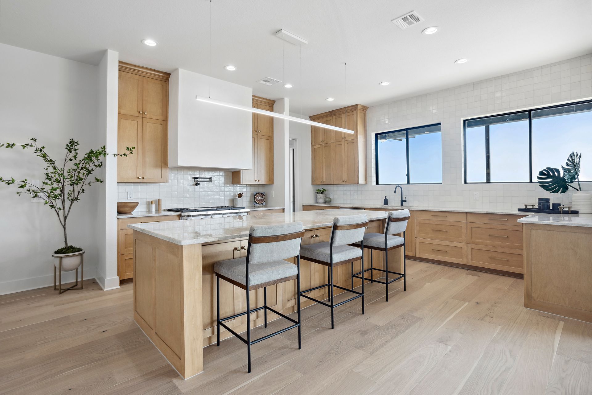 Bright modern kitchen with light wood cabinets, white countertops, island stools, and large windows