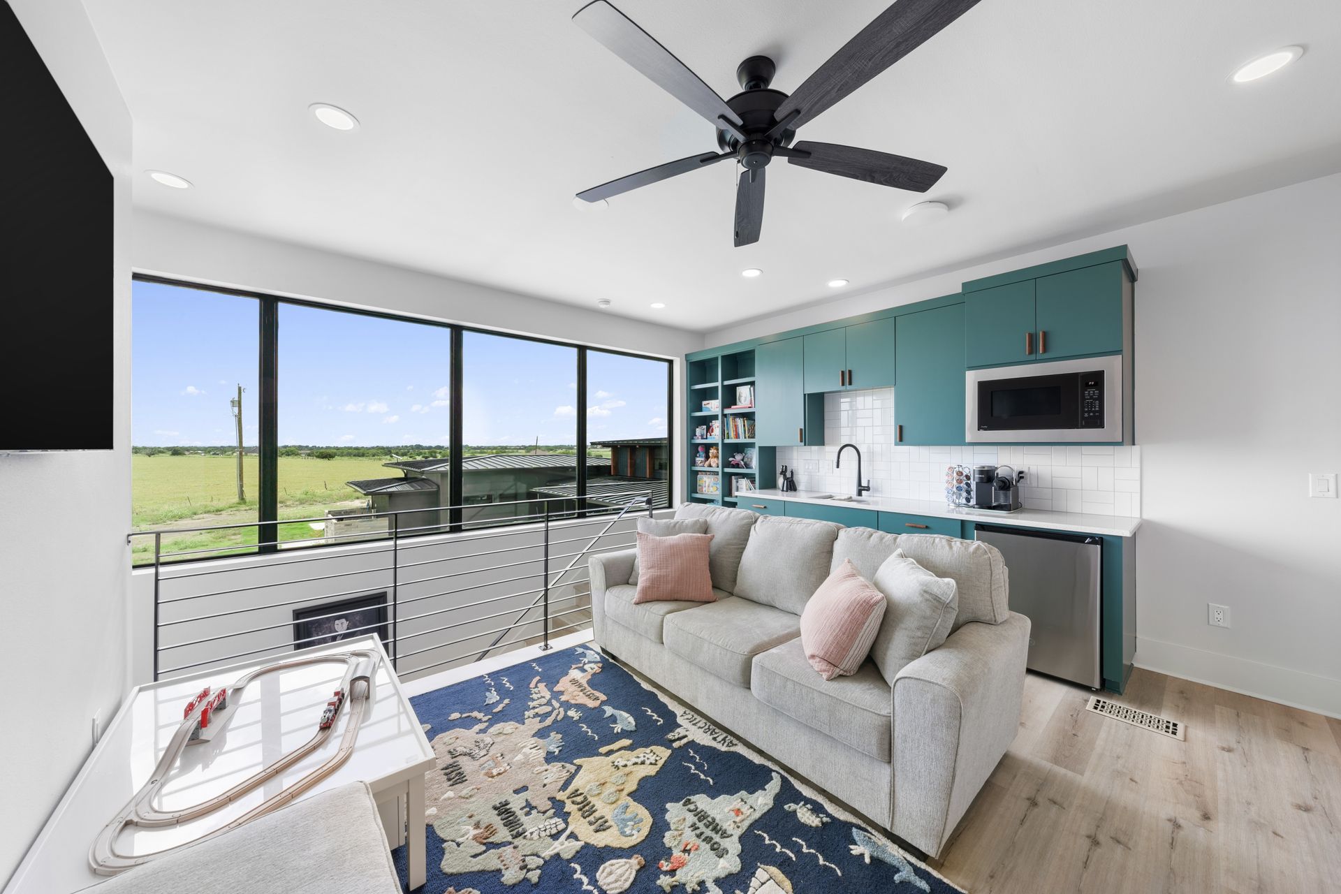 Bright modern living room with teal kitchen, large windows, gray sofa, and ceiling fan