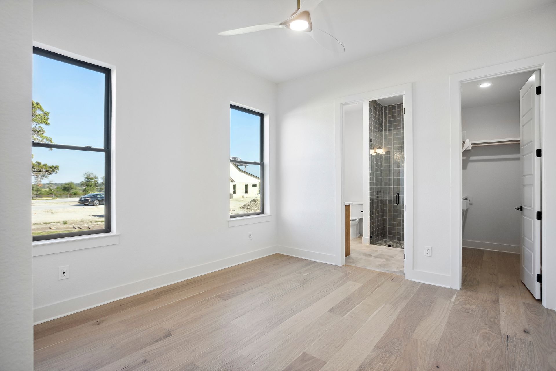 Bright empty bedroom with white walls, wood floors, windows, and an en suite closet/bath area