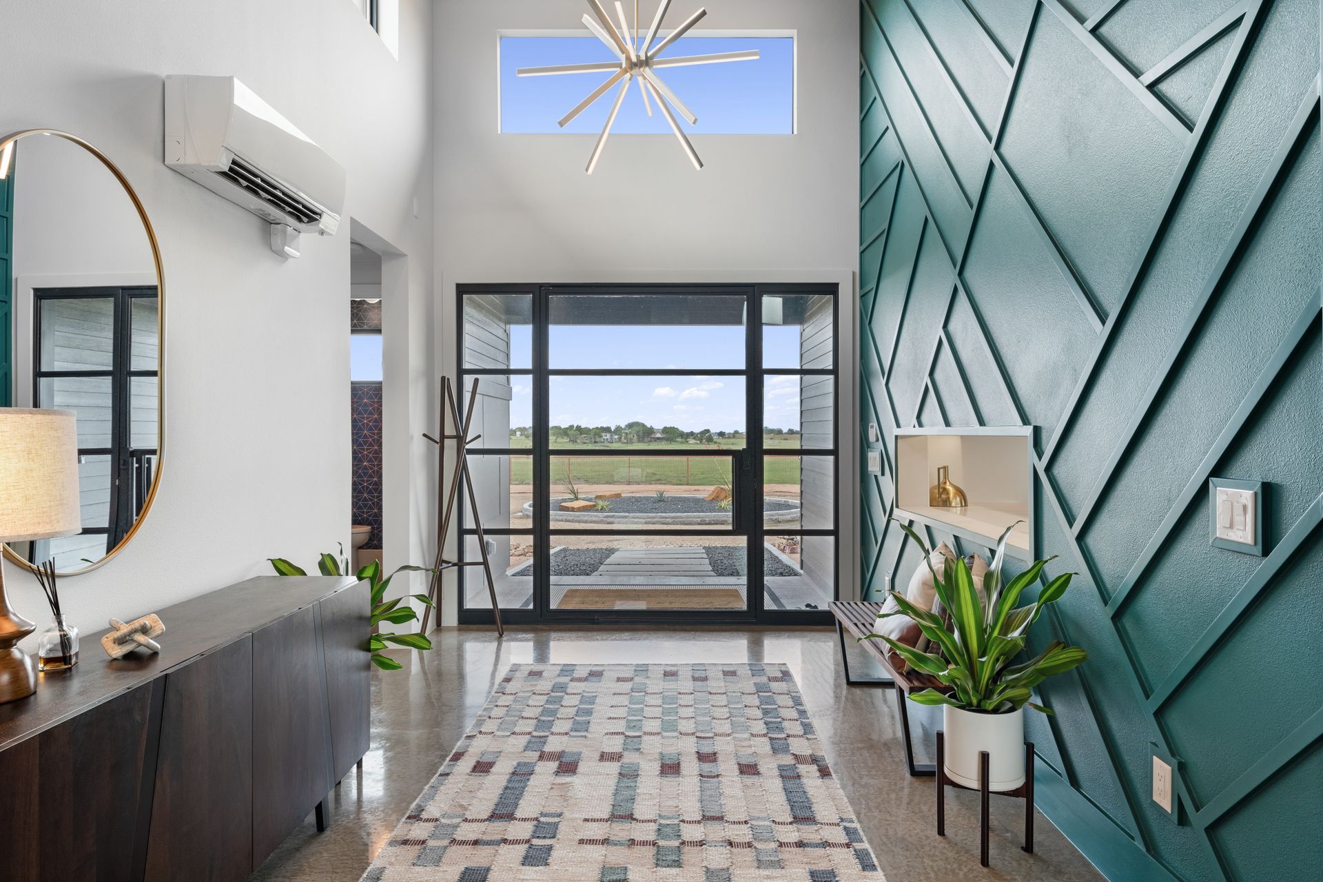 Modern entryway with teal geometric wall, dark console, round mirror, plant, and large front window.