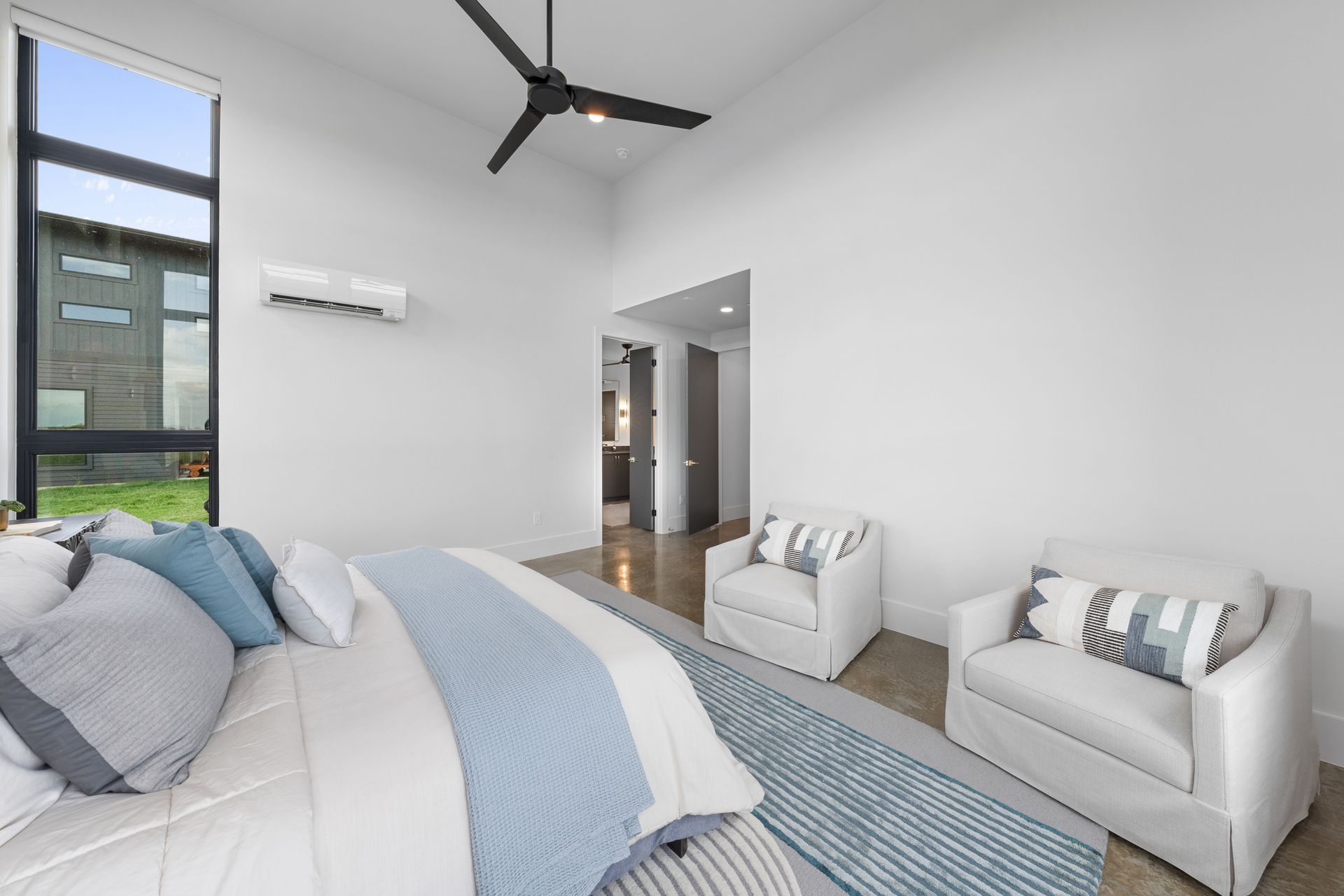 Bright modern bedroom with white walls, blue accents, large window, ceiling fan, and seating area