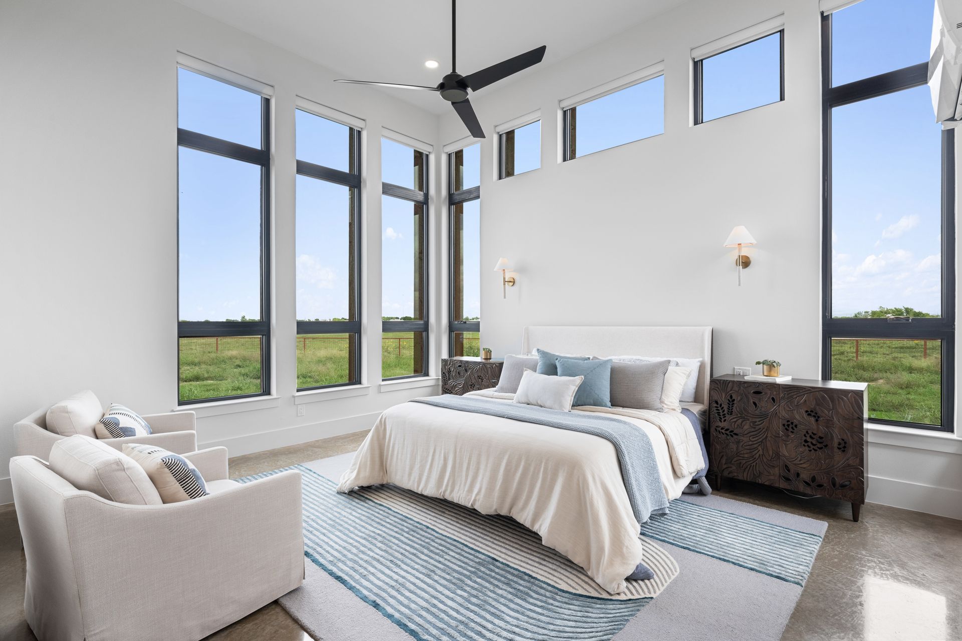 Bright modern bedroom with large windows, white bedding, and a light armchair overlooking green fields