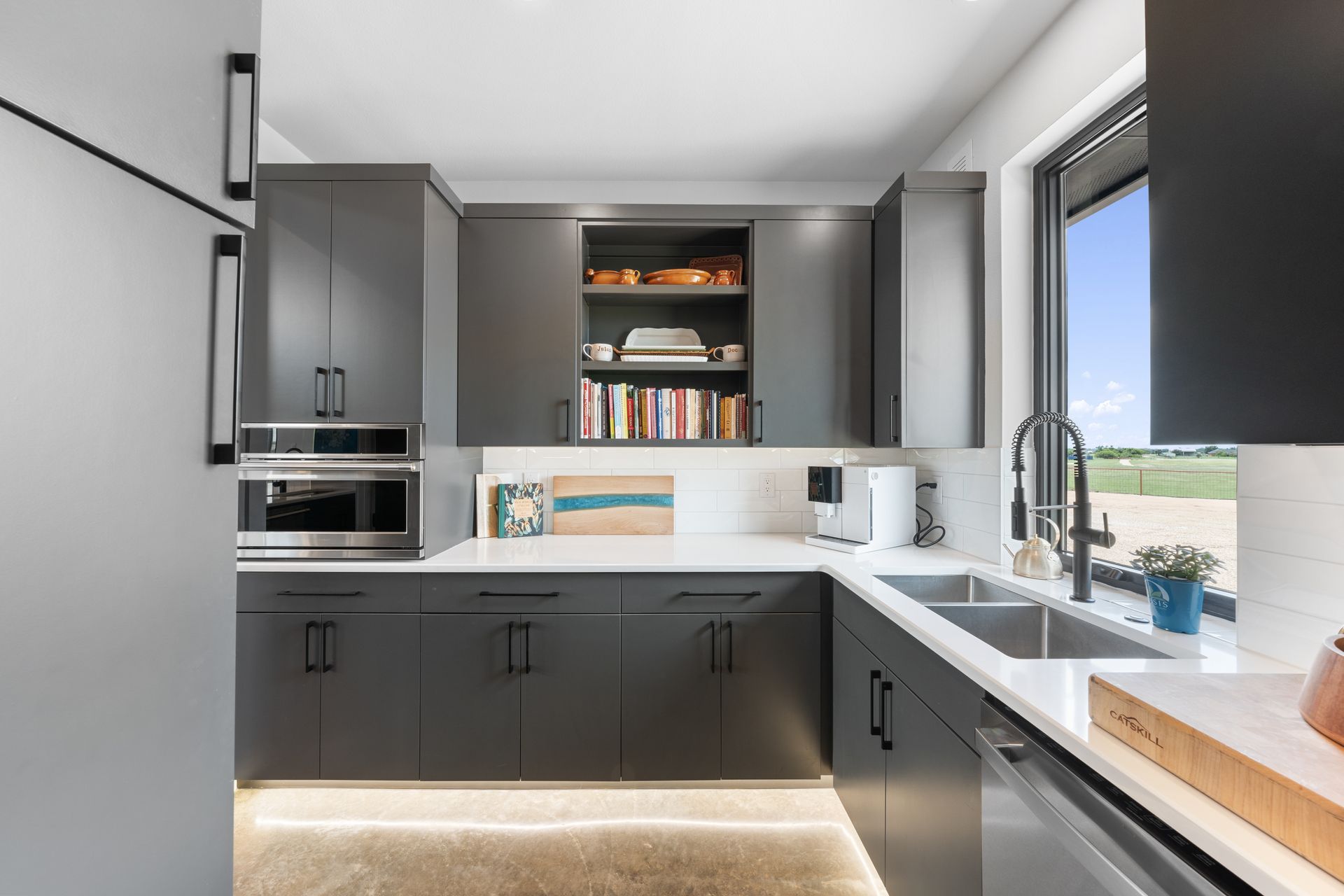 Modern gray kitchen with white countertops, built-in appliances, and a window view of a patio and sky