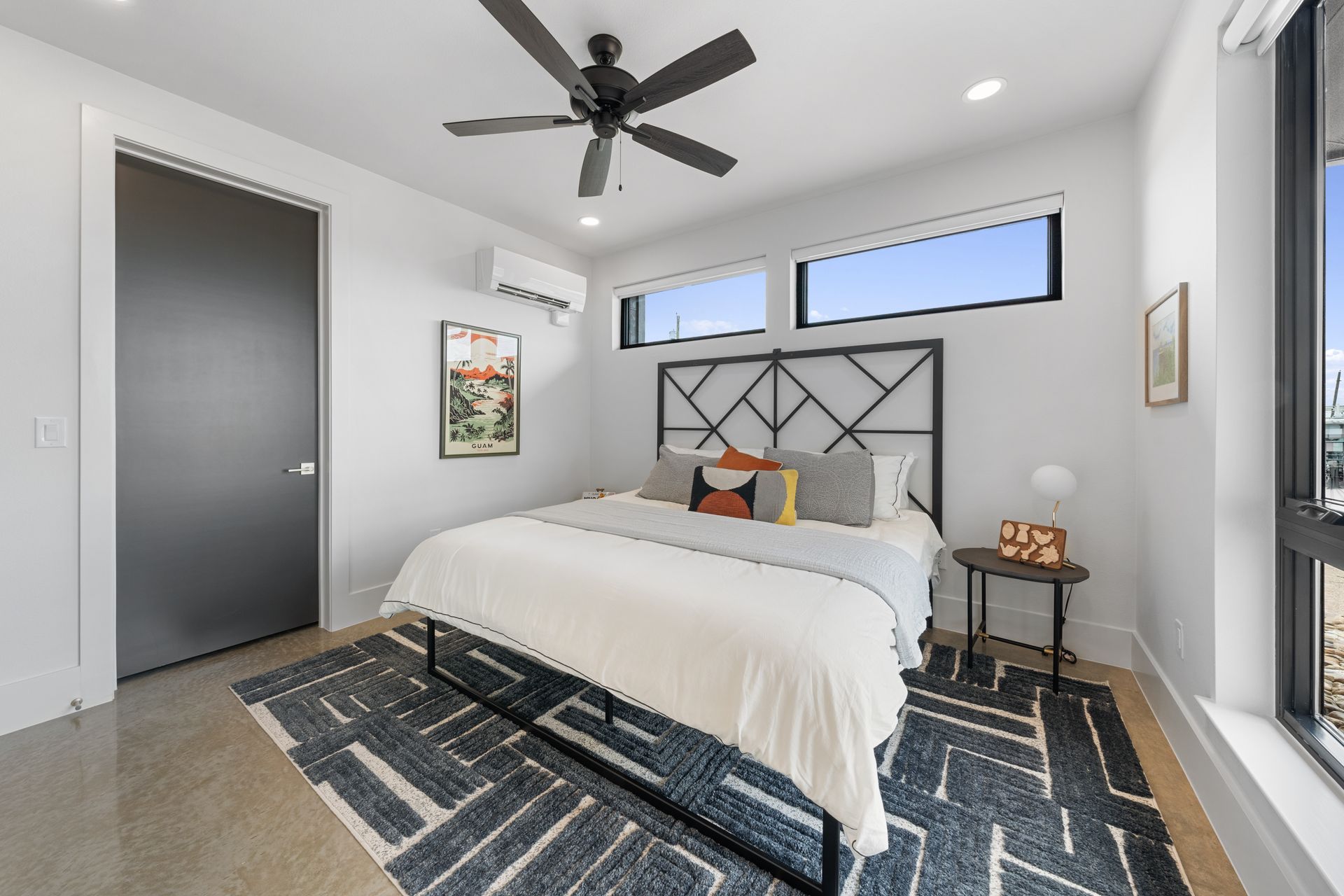 Modern bedroom with a black bed frame, gray walls, patterned rug, and ceiling fan, lit by natural light.